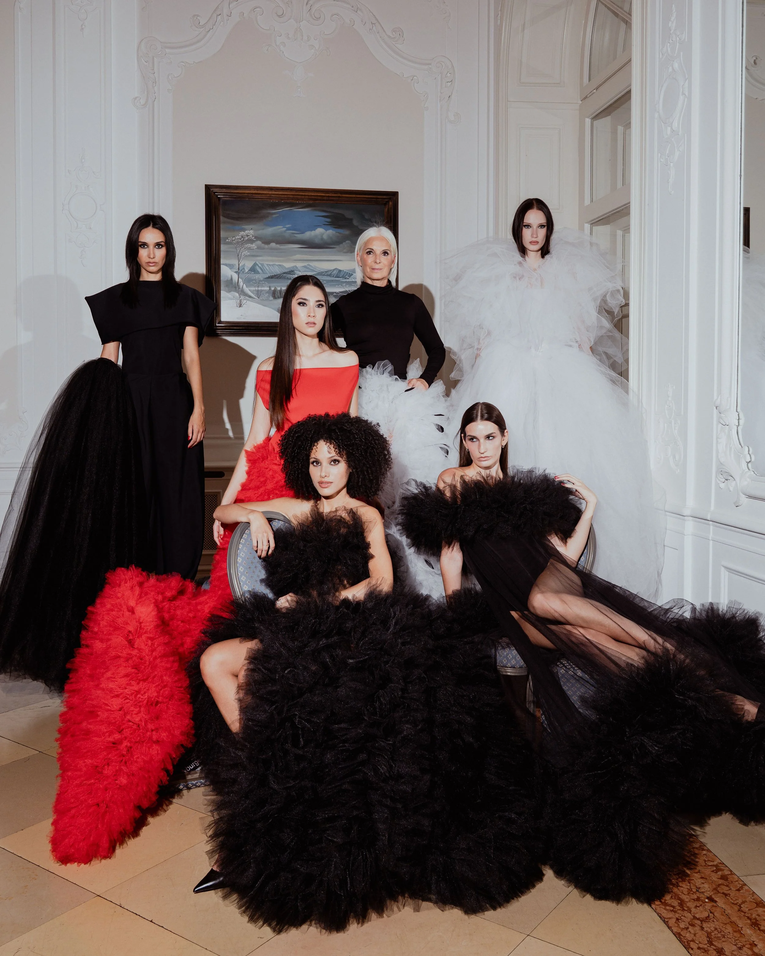 A group of seven women dressed in elaborate fashion, with some wearing feathered clothing, pose in a room with ornate white walls and a painting of a snowy landscape in the background.