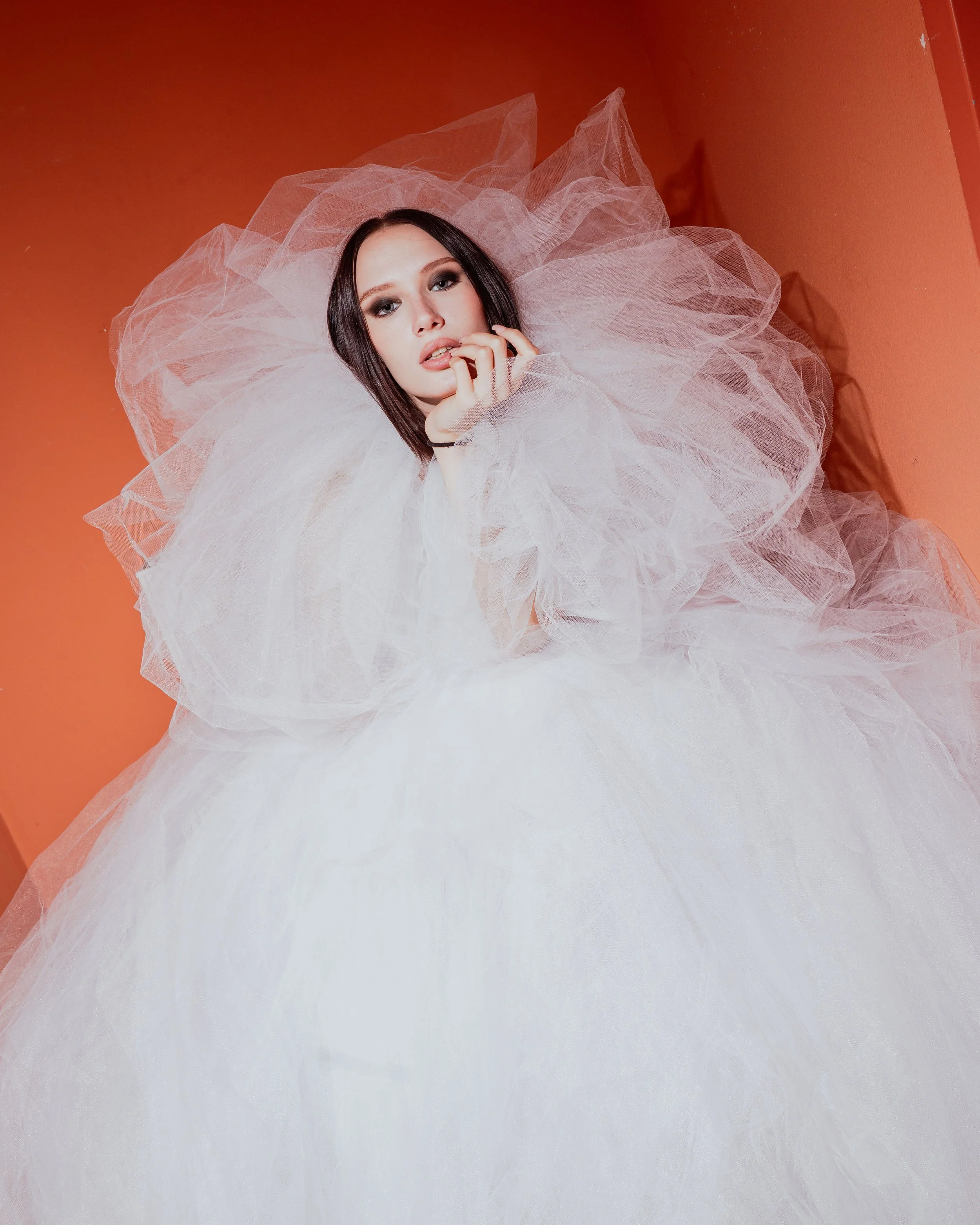 A woman with dark hair and makeup surrounded by pink tulle fabric, sitting against an orange wall.