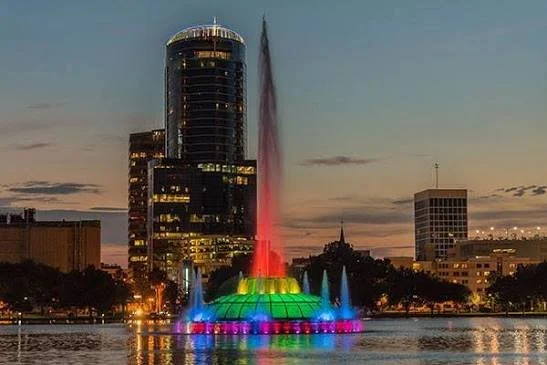Lake Eola fountain illuminated in vibrant rainbow LED colors reflecting across dark water at night against the Downtown Orlando skyline