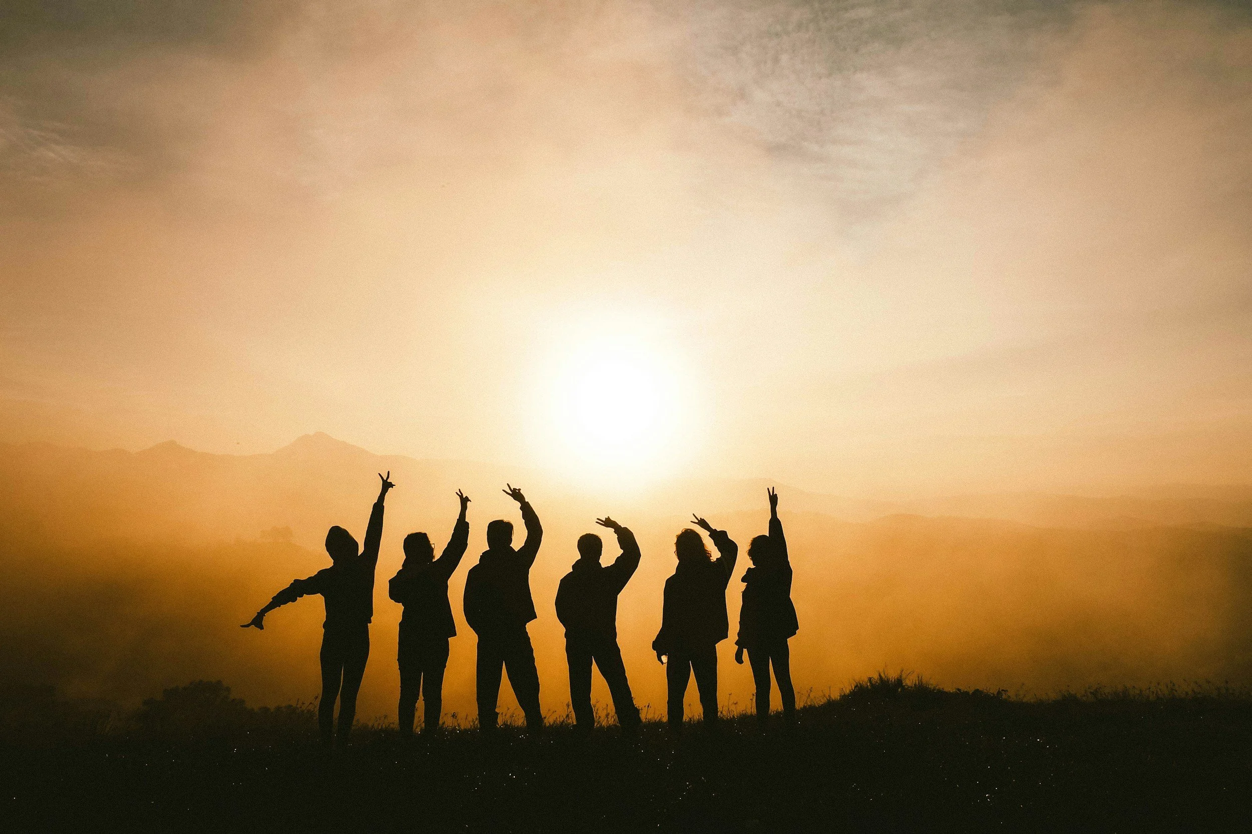 Silhouettes of a diverse group of people raising their hands in peace signs against a sunset over mountains, representing community healing at Calming Tides Therapy.