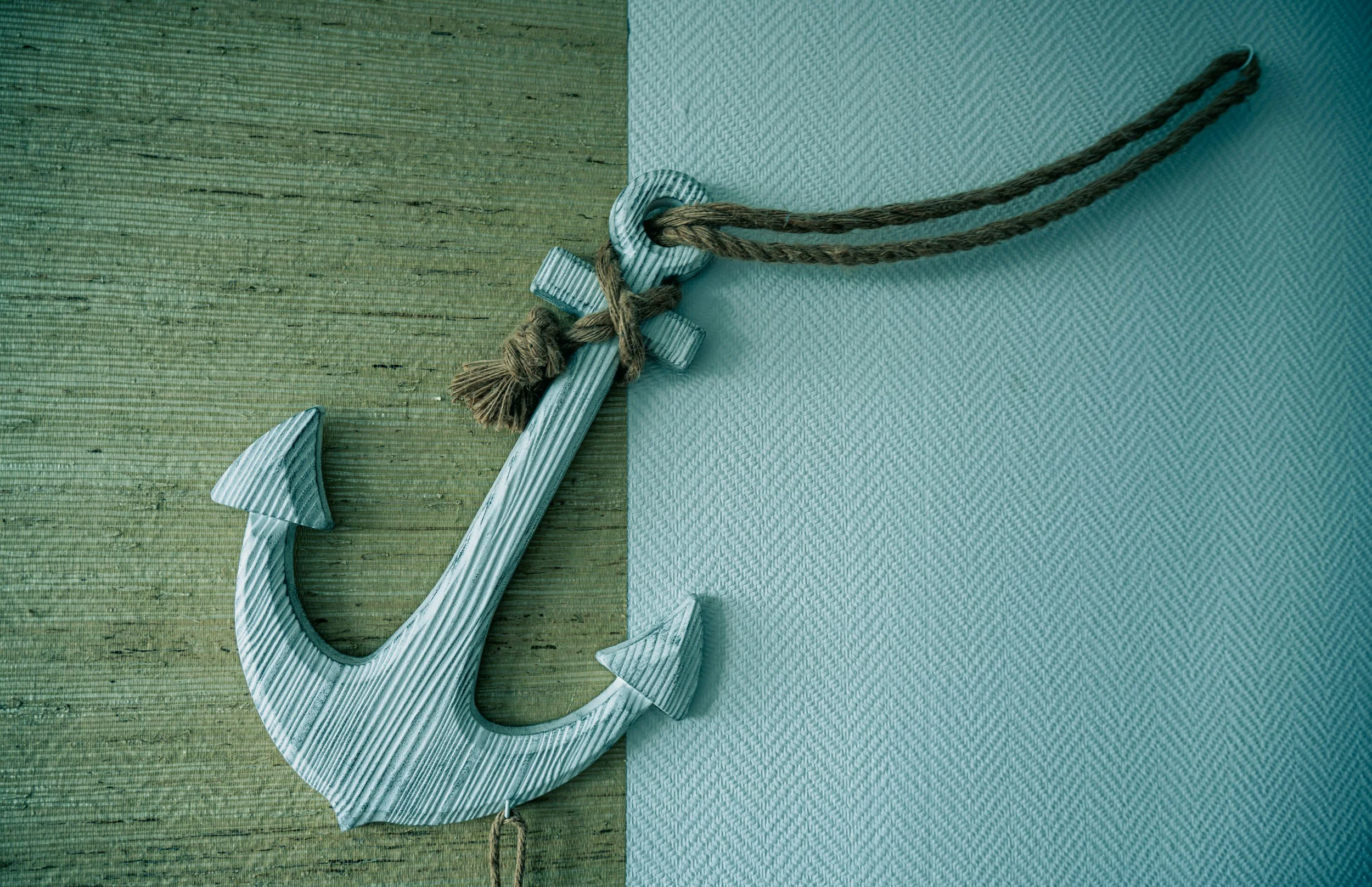 Close-up of a large white anchor with a textured surface, tied with a brown rope, resting on a wooden surface next to a piece of light blue fabric.