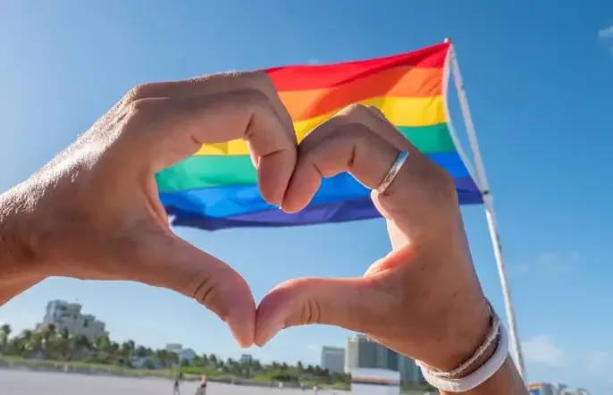 Two hands forming a heart symbol against a brightly lit rainbow pride flag, representing love, inclusivity, and LGBTQ+ community support.