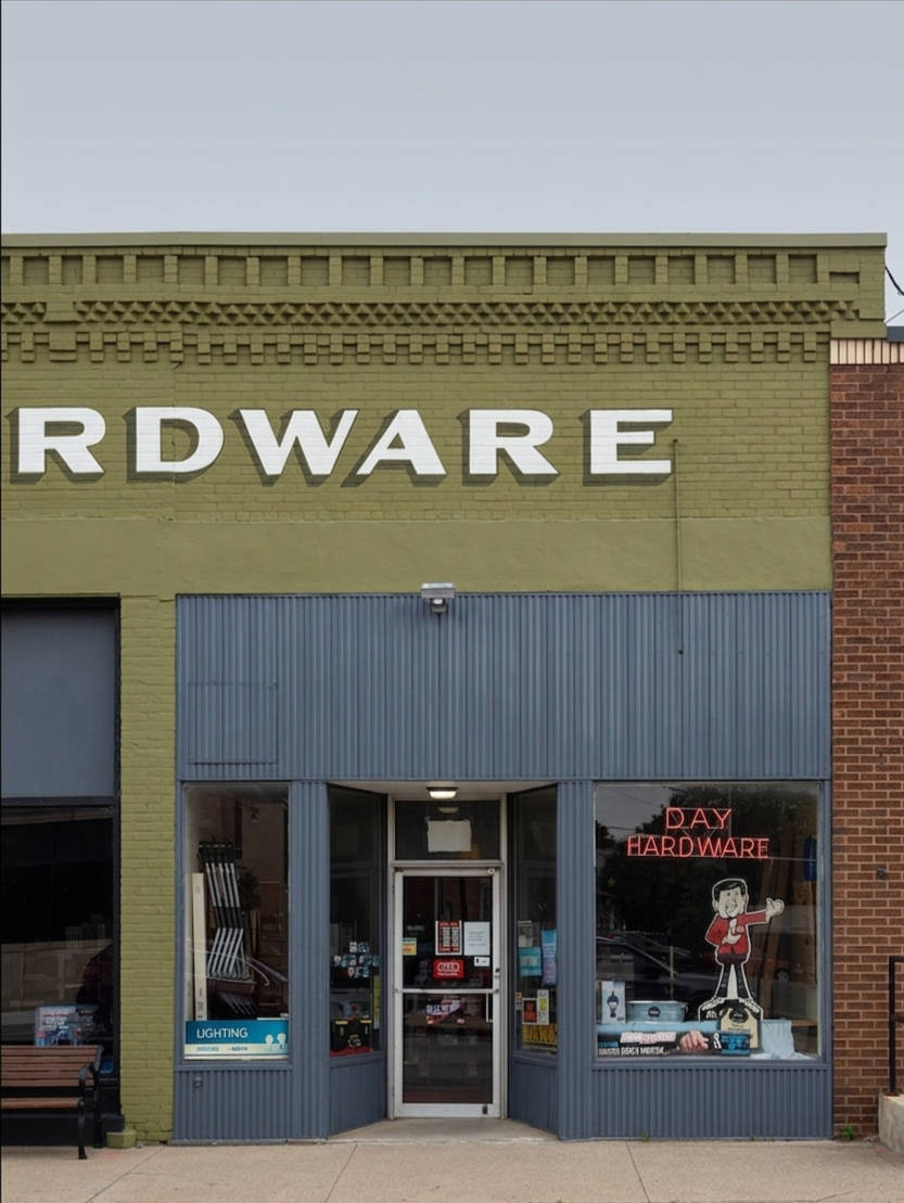Front view of a hardware store with a sign that reads 'HARDWARE' partially visible on the building. The shop has a glass door and large windows displaying hardware supplies, a neon sign that says 'DAY HARDWARE', and a cartoon character window decal. The store is painted green and gray with brickwork on the right side.