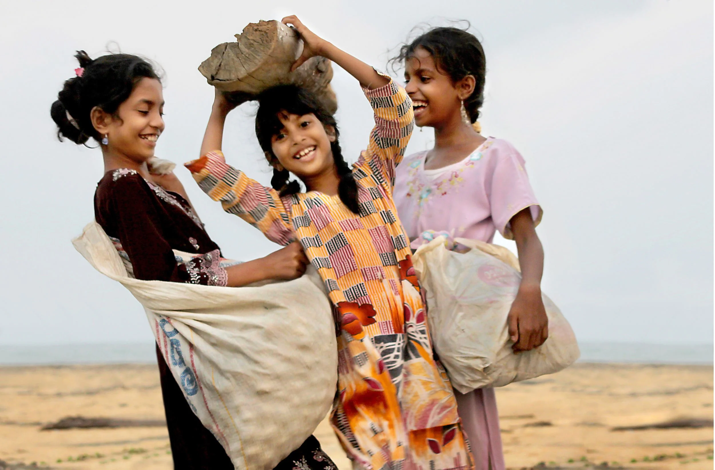 Three young girls on the beach smiling and holding a large piece of driftwood between them.