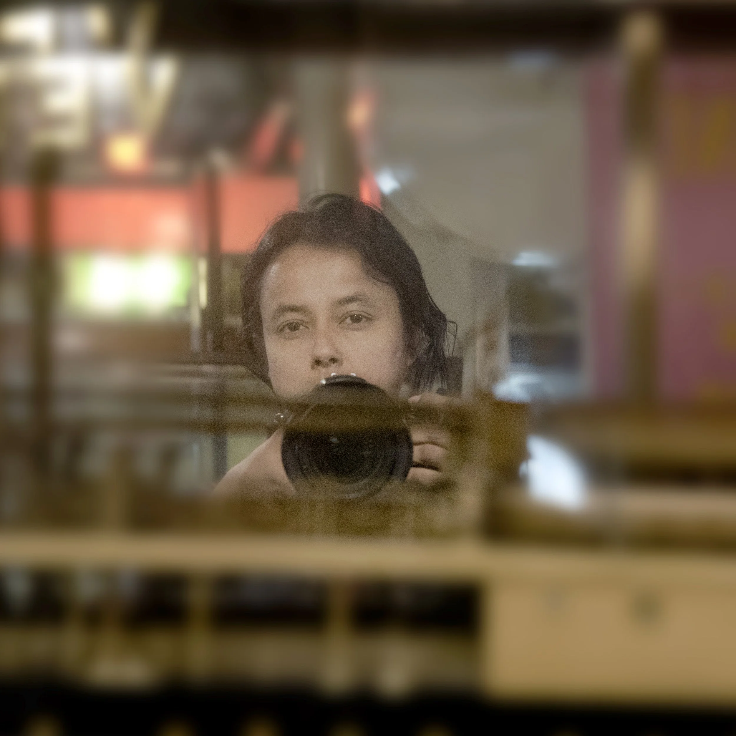 A young woman with dark hair taking a selfie in a mirror, with blurred lights and indoor setting in the background.