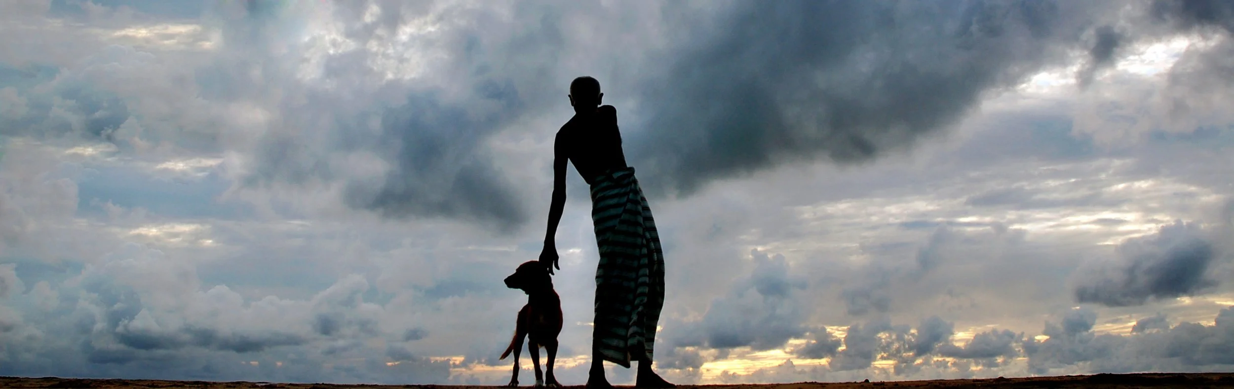 Two figures move across the shore beneath a heavy sky, held for a moment between weather, silence, and home.
