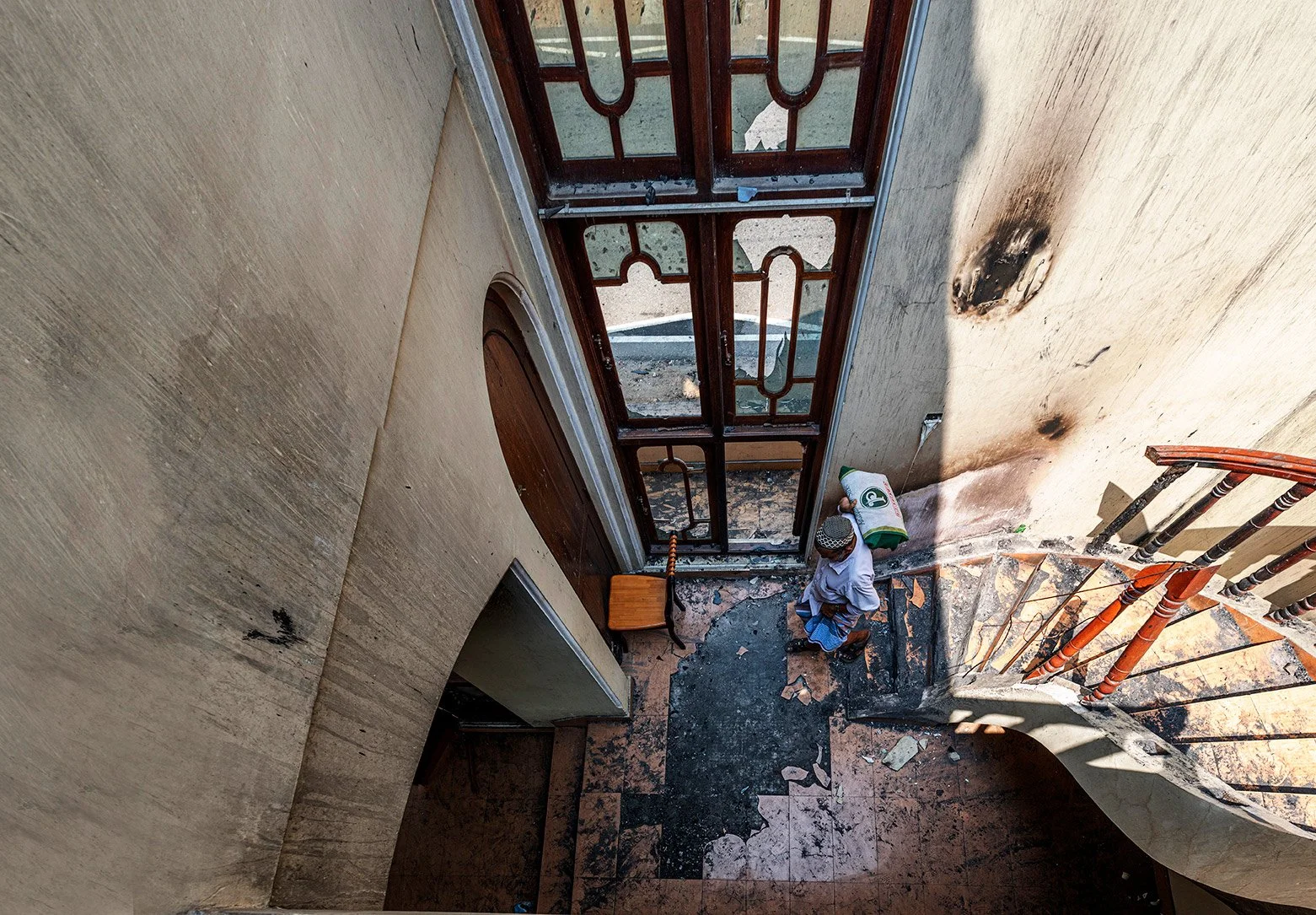 A man carrying a bag over his shoulder stands inside a building near the entrance, which shows signs of fire damage. The floor is charred and debris is scattered. The staircase next to him has a wooden banister, and the wall near the entryway has black smoke marks.