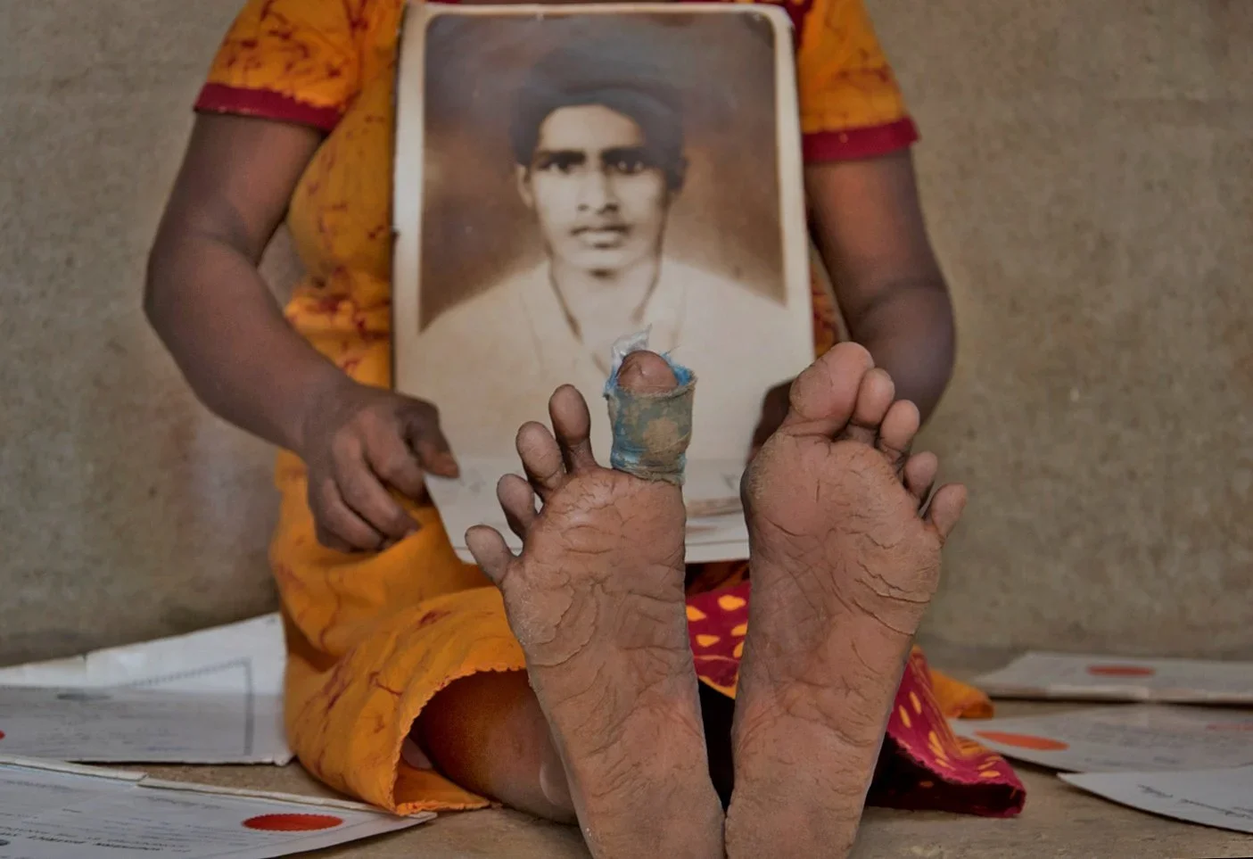 A person wearing a yellow and red outfit sitting on the ground, holding a framed black and white portrait of a young man. In front of them, their feet are visible, showing cracked and dry skin, with one toe wrapped in cloth.