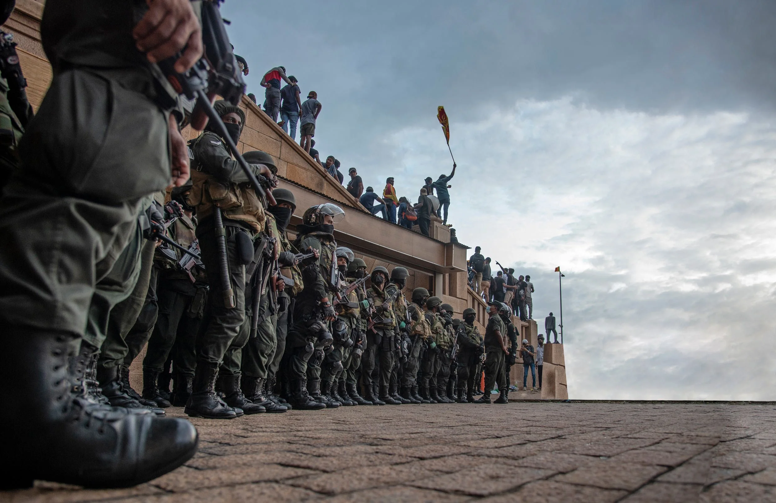 Line of police officers in riot gear standing outside on a paved area, facing forward. Several people are gathered on the ledge above them, some holding flags, with a cloudy sky in the background.