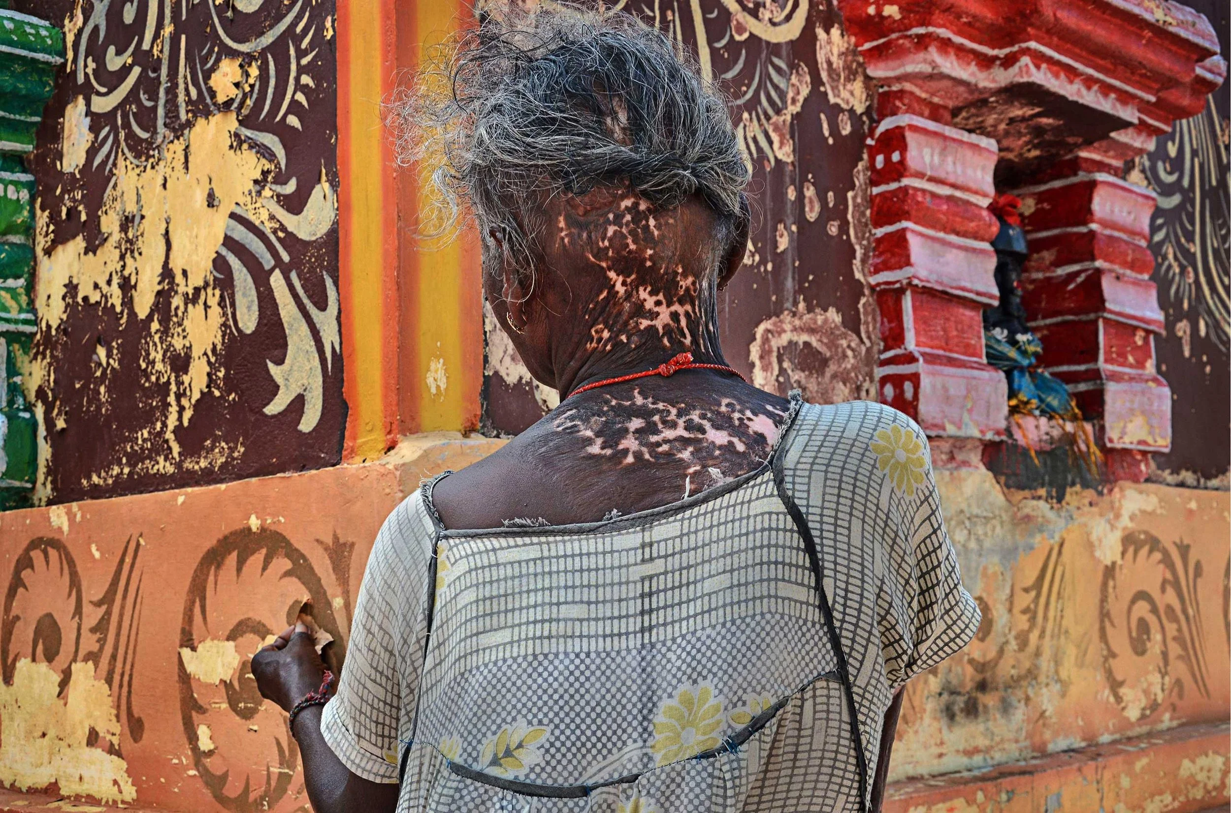 A woman stands against a weathered wall, where colour, age, and survival sit quietly in the same frame.
