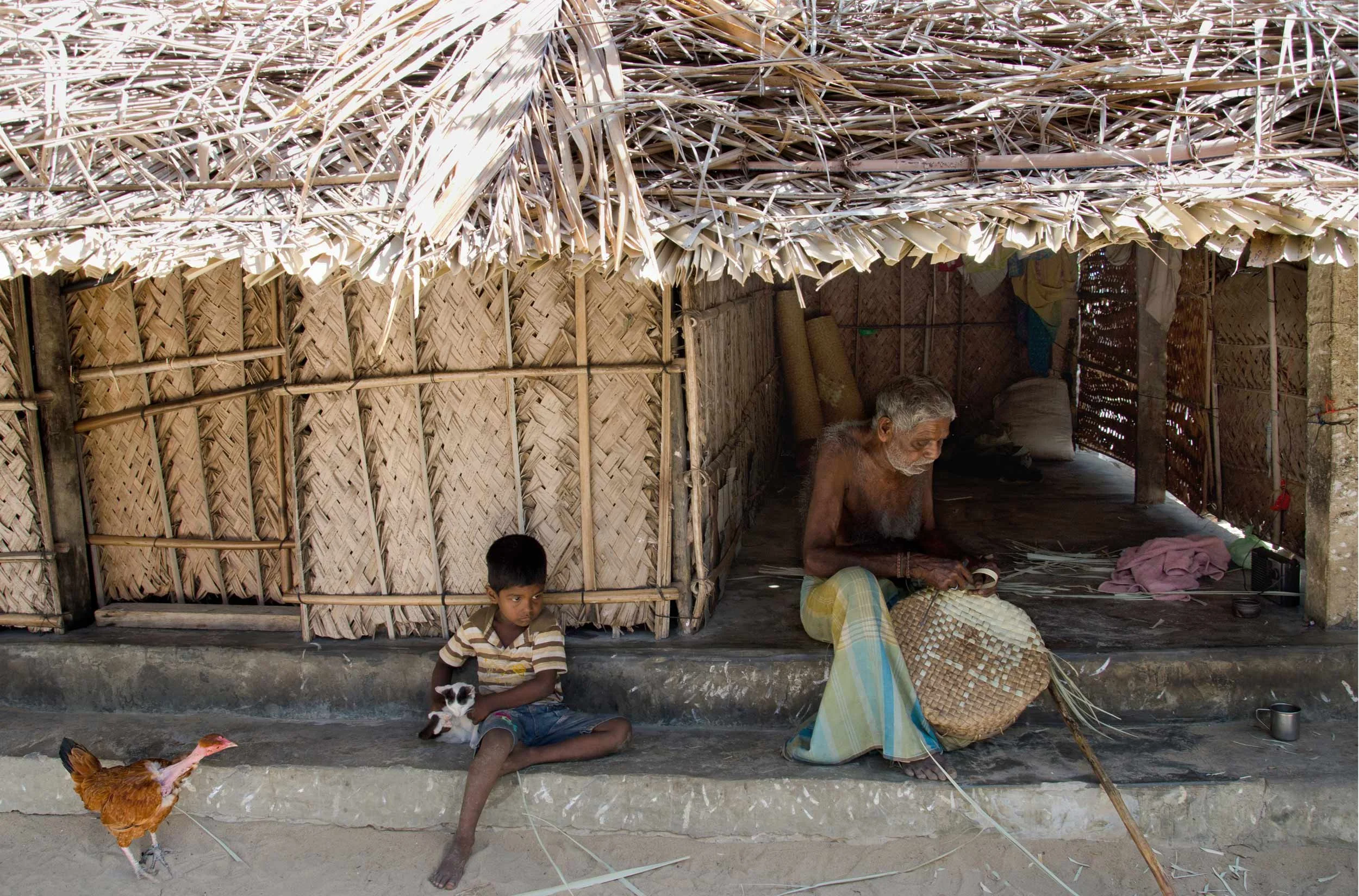 An elderly woman and a child sit inside a woven shelter, where ordinary life continues in the long shadow of war.