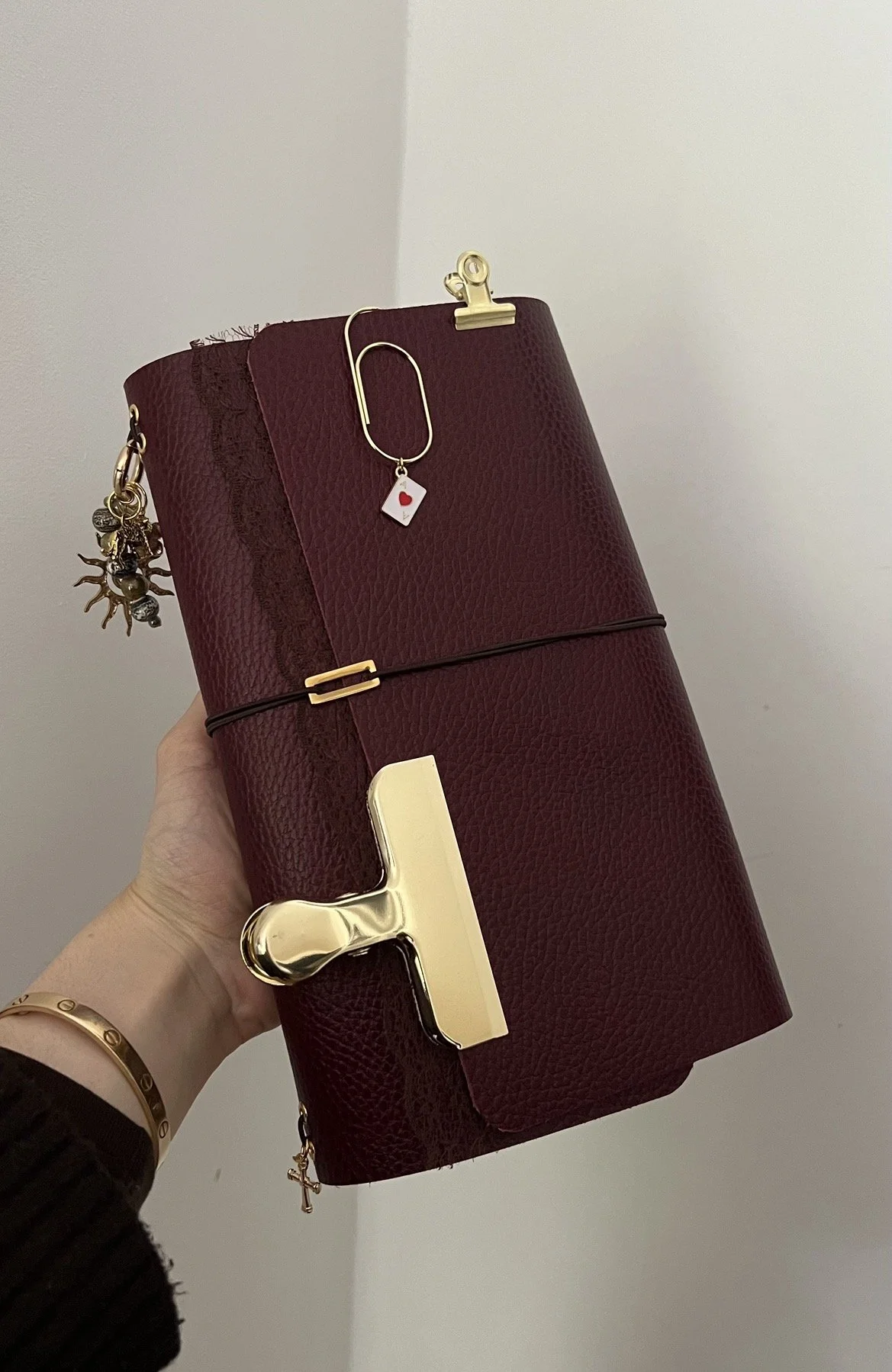 A burgundy textured notebook held in hand with Gold and black clip, gold safety pin with a small white card with a red heart, small charms hanging from the side, and a gold pen with a large clip.