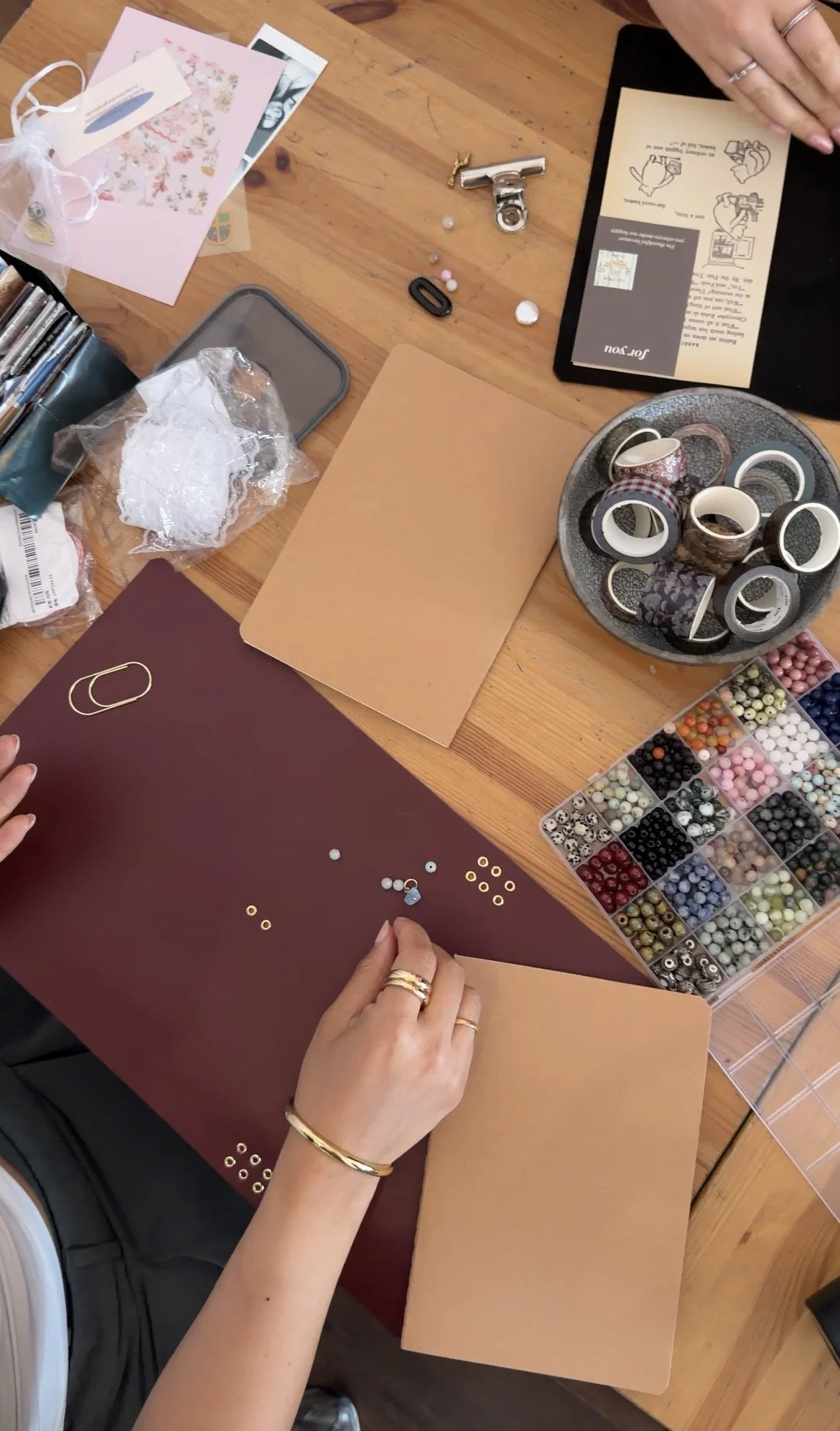 Person preparing to decorate a scrapbook with beads, washi tapes, and stickers on a wooden table.