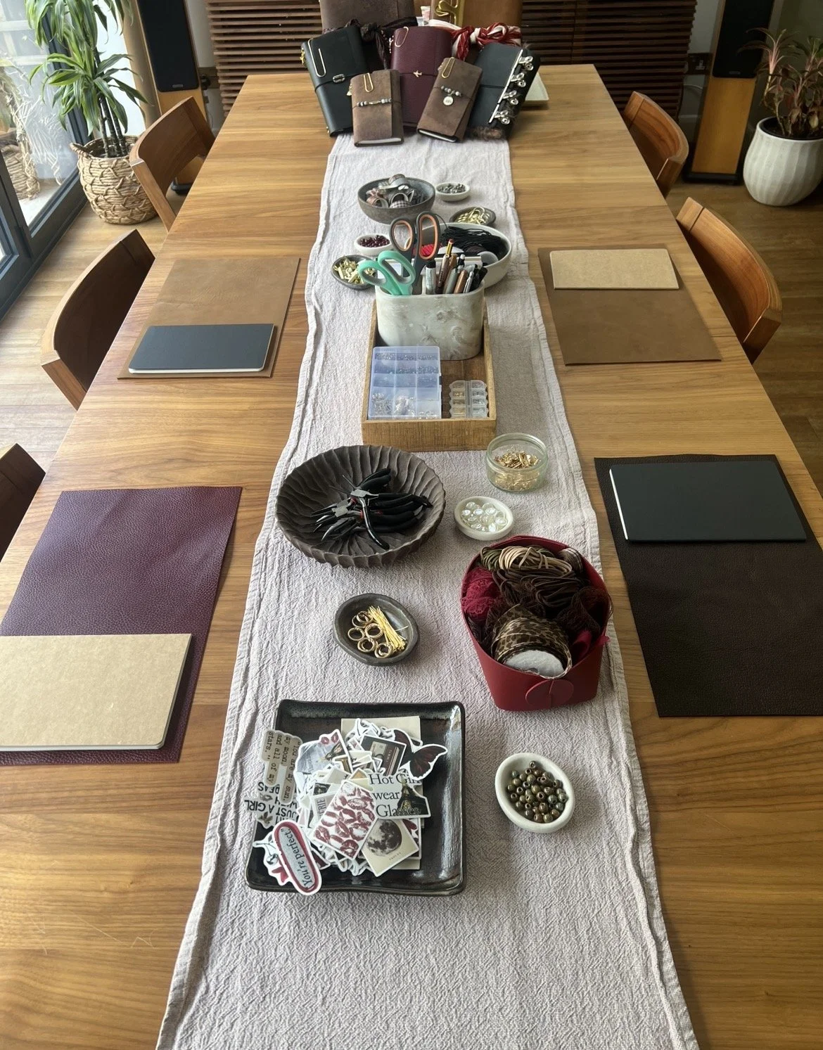 A long wooden table with a beige table runner, surrounded by wooden chairs. The table holds various craft and office supplies, including scissors, pins, jewelry findings, buttons, embellishments, and decorative items. There are notebooks and folders 