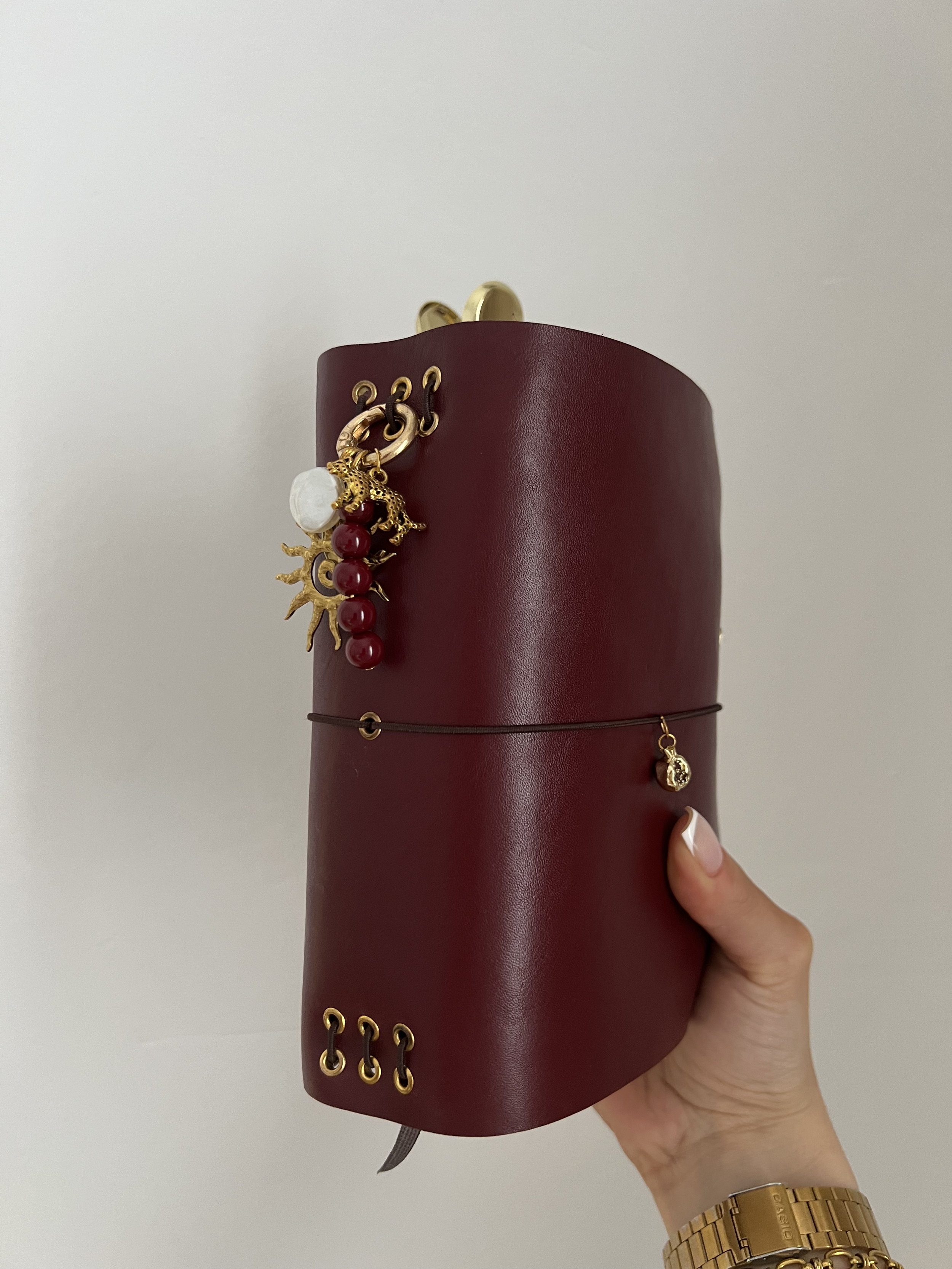 A hand holding a cylindrical red leather jewelry box decorated with gold eyelets and charms, with one large gold and white charm attached to the front.