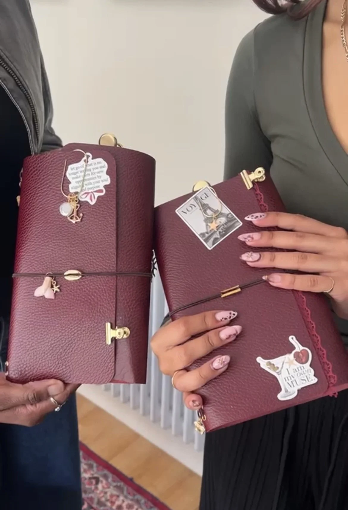 Two women holding burgundy leather wallets with gold accents and charms. One wallet has a tag with a palm tree and a woman in a dress, the other has a tag with an Eiffel Tower and the word "LOVE." The women are showing off the wallets in a well-lit r