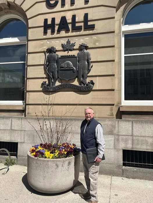 Bob standing in front of Brantford city hall building with a large flower pot and a crest above him.