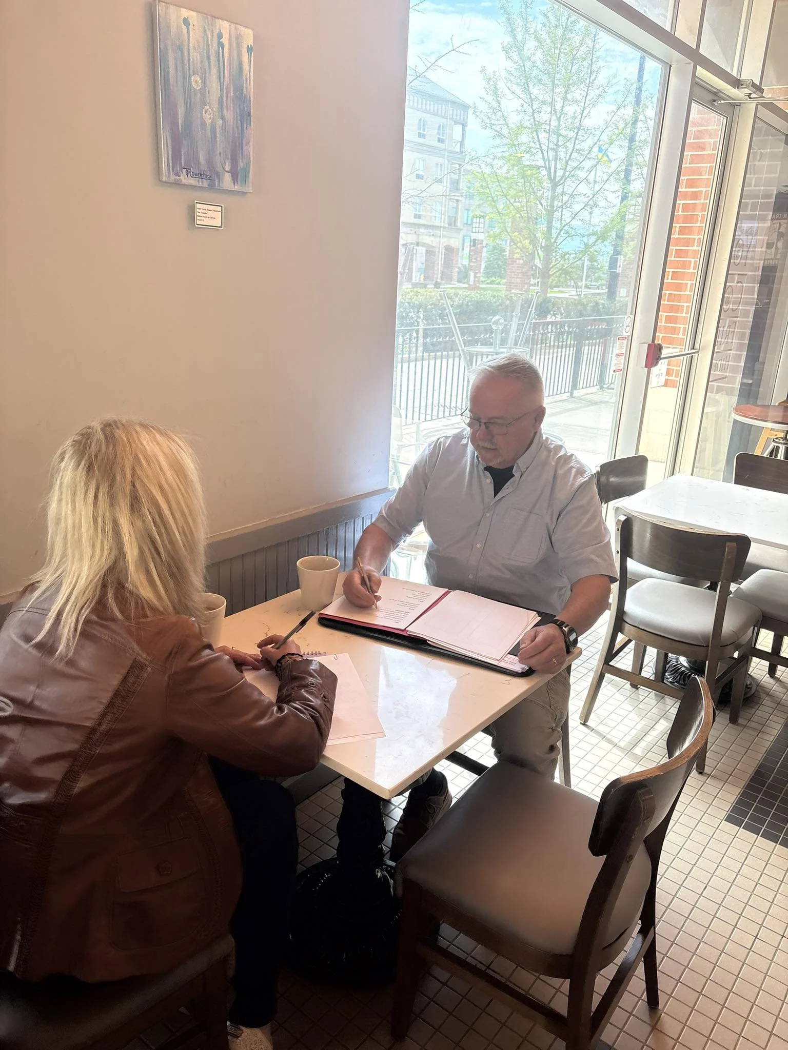 A man and a woman sitting at a table in a cafe, engaged in a discussion. The man is writing in a notebook, and the woman is holding a pen. The cafe has large windows with a view of the outdoors, including trees and buildings. There are cups on the ta