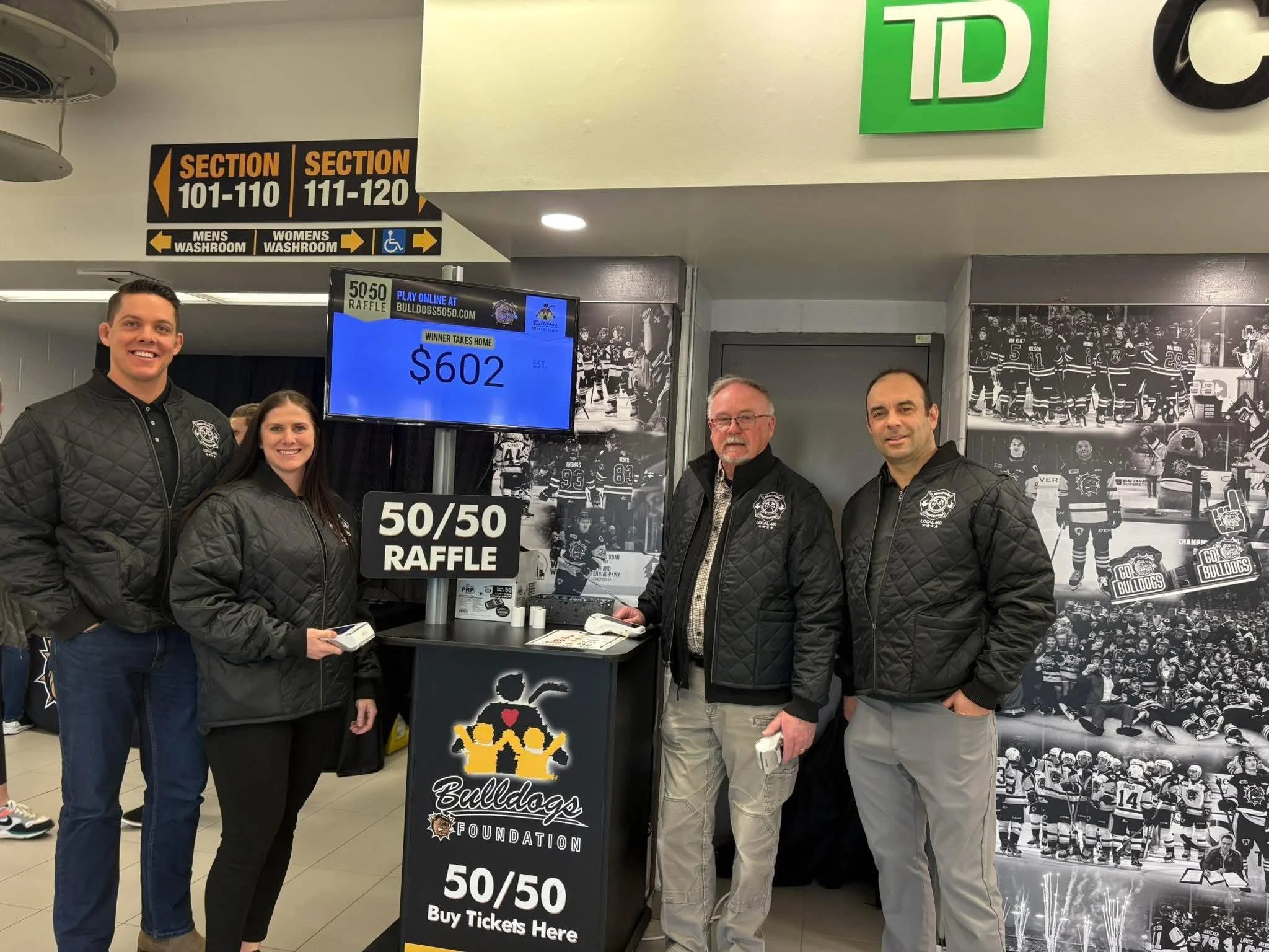 Four people standing behind a ticket booth for a hockey game with black and white photos of hockey players in the background. They are wearing black jackets, some with a logo, and smiling at the camera. There is a sign in front of them for a 50/50 ra