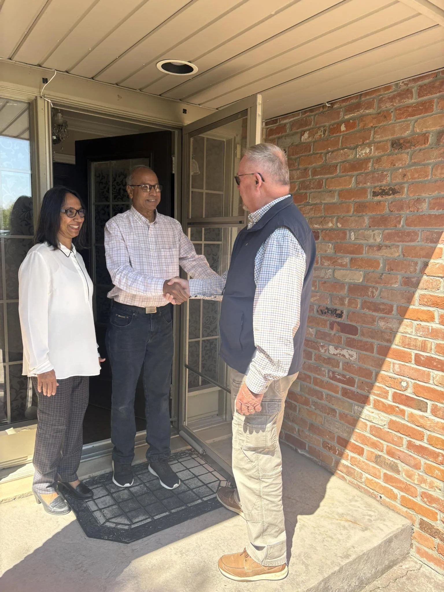 Bob and neighbours standing outside near a house entrance, shaking hands and smiling. One woman and two men, with a brick wall and doorway in the background.
