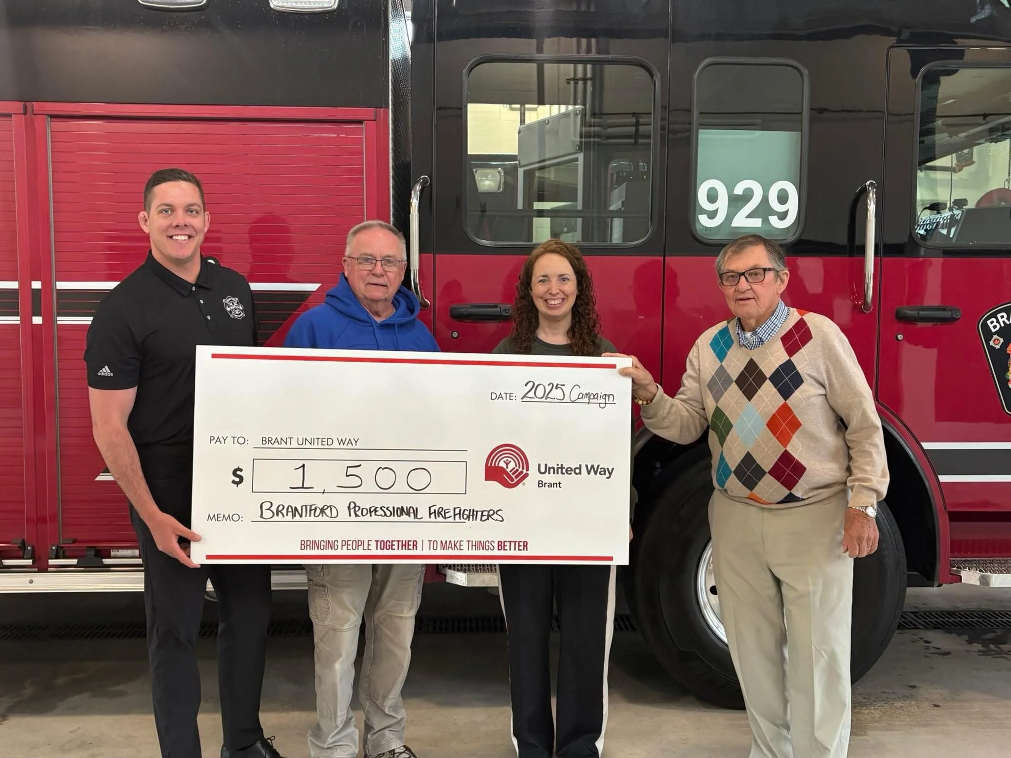 Bob and his team standing in front of a fire truck, holding a large check made out to Brant United Way for $1,500, for a 2025 campaign, with a woman and a man with glasses holding the check.