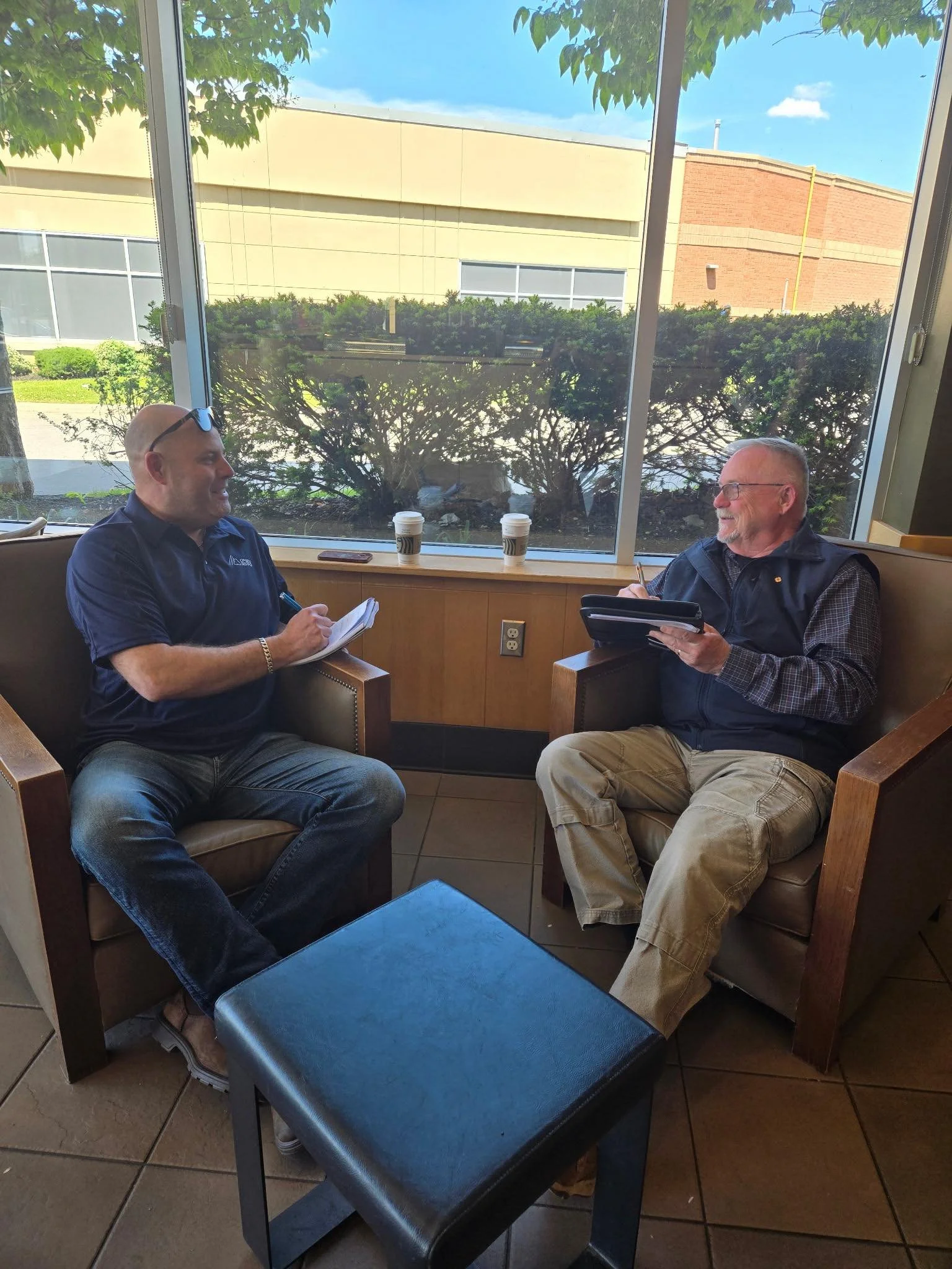 Bob with a man sitting in a coffee shop, talking with each other. Both are holding notepads and pens, and are smiling. There are two coffee cups on the windowsill behind them, and the window shows bushes and a building outside.