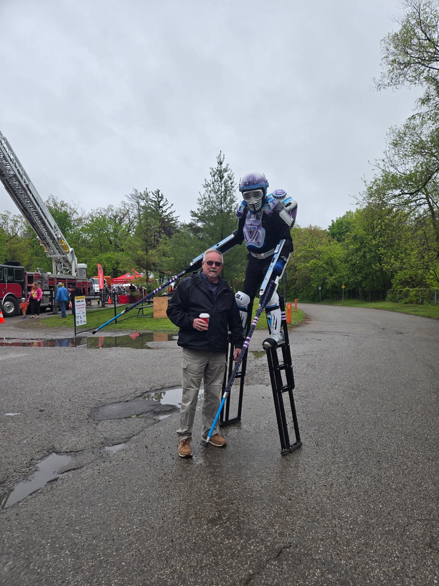 Bob holding a coffee cup, standing next to a large humanoid robot on stilts at an outdoor event. There are fire trucks, tents, and people in the background, with cloudy weather and green trees.