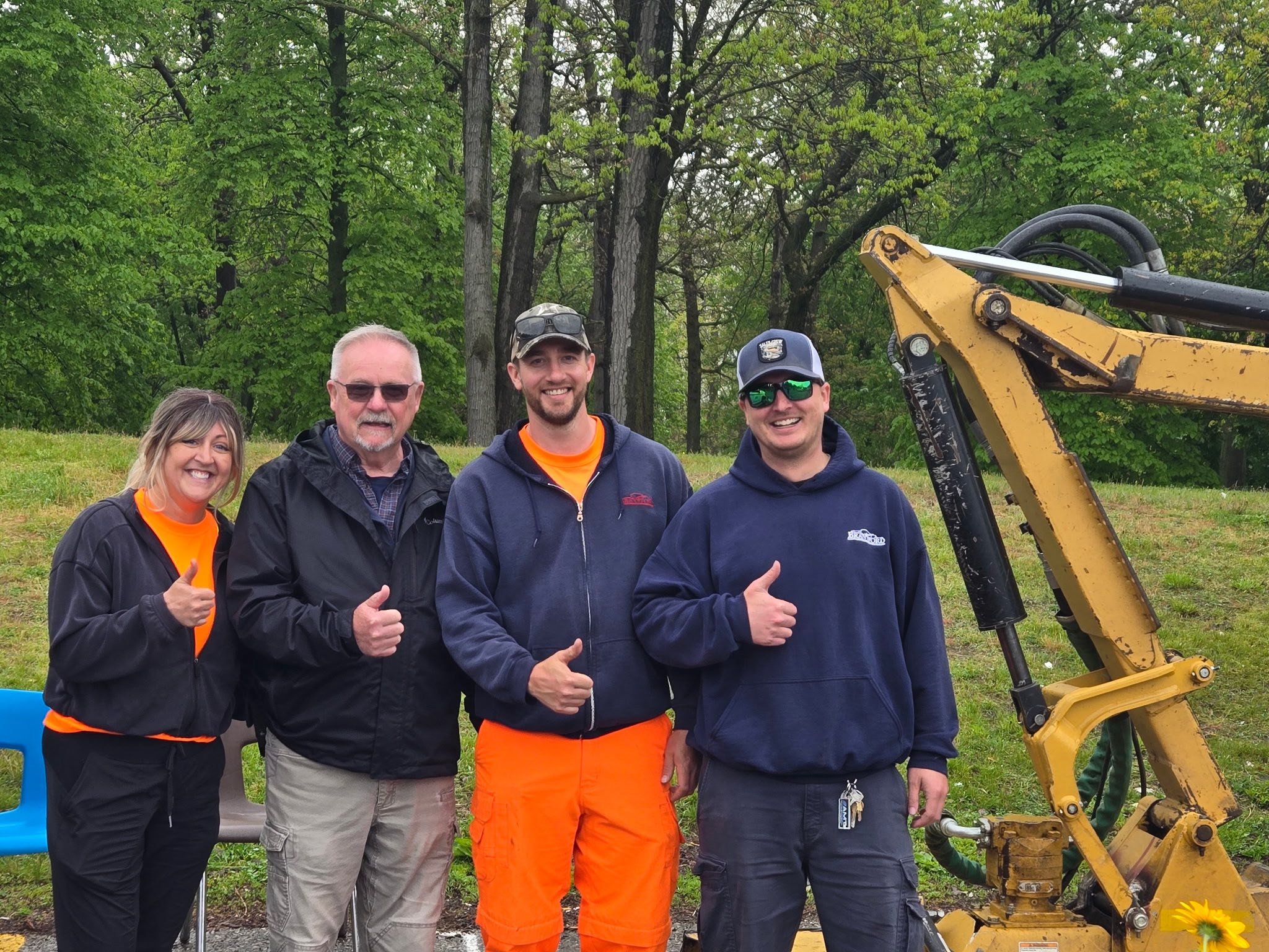 Bob and a group of five smiling people outdoors standing next to a yellow construction machine, with green trees in the background.