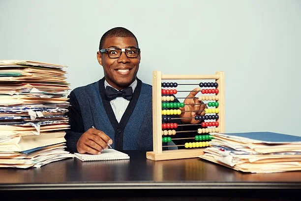A man with glasses, wearing a bow tie and sweater, sitting at a desk with piles of papers on both sides, smiling, with an abacus in front of him.