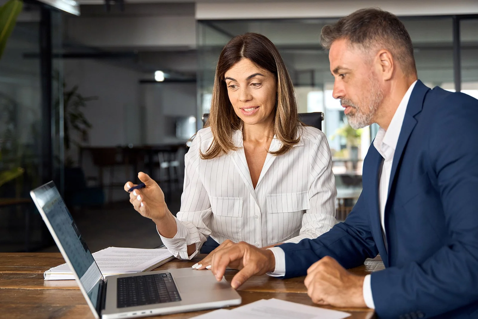 Two business professionals, a woman and a man, sit at a table looking at a laptop screen and discussing in an office setting.