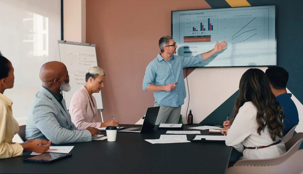 A man in a blue shirt presenting data on a large screen to a group of five people sitting around a conference table, with charts and graphs displayed.