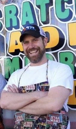 A man with a beard and a big smile, wearing a baseball cap and an apron, standing in front of a colorful banner with text.