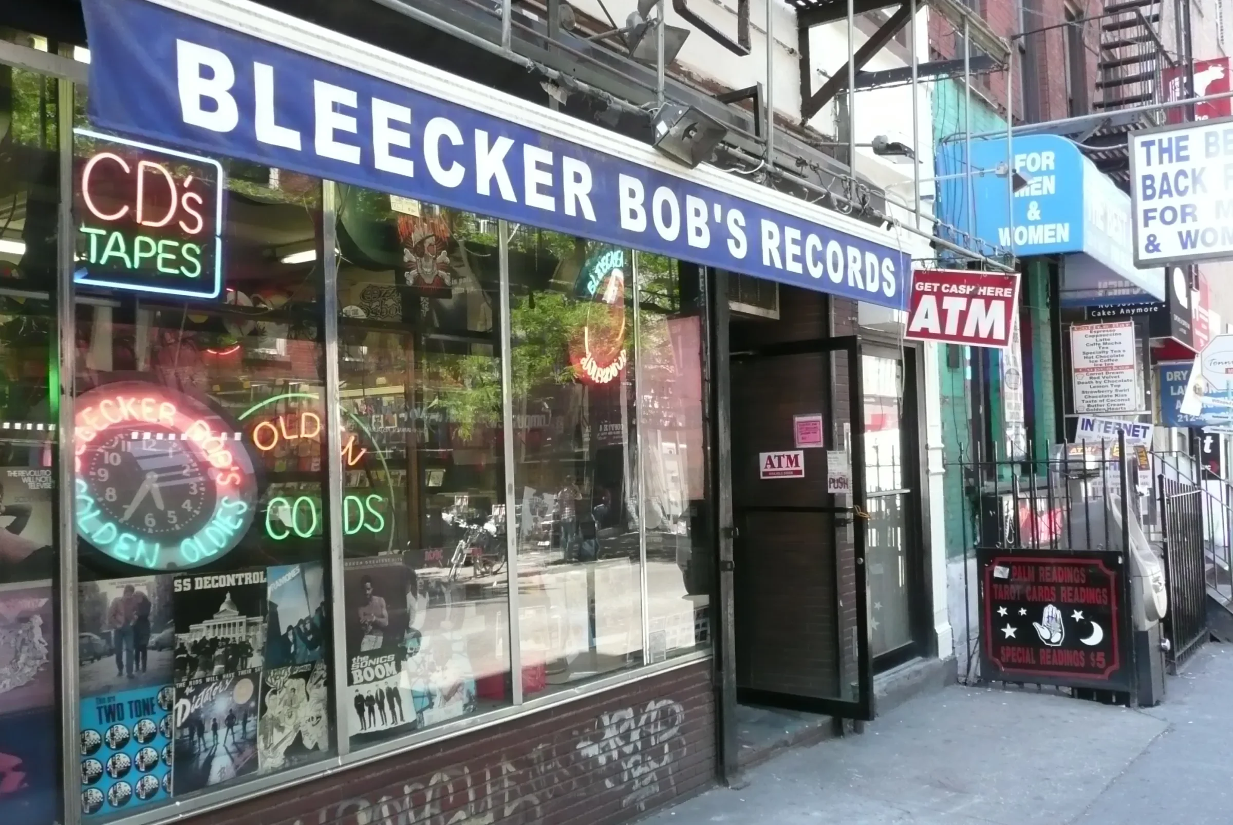 Storefront with a large blue sign that reads 'Bleecker Bob's Records'. The window display has colorful neon signs advertising CDs, tapes, old records, and retro music posters. There is a door labeled 'ATM' next to the entrance and a sign on the sidewalk advertising tarot card readings for $5.