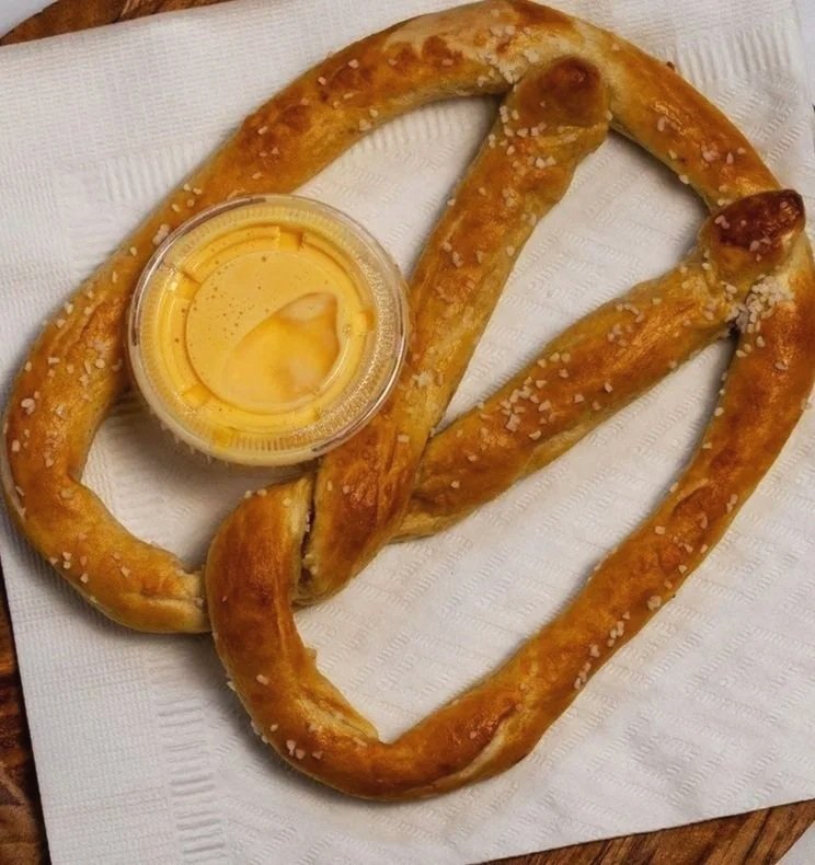 A large pretzel with coarse salt crystals, accompanied by a small cup of mustard on a white napkin.