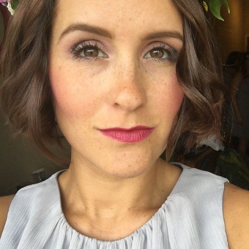 A woman with short, wavy brown hair and light freckles, wearing a sleeveless light gray top, and lipstick, posing indoors.