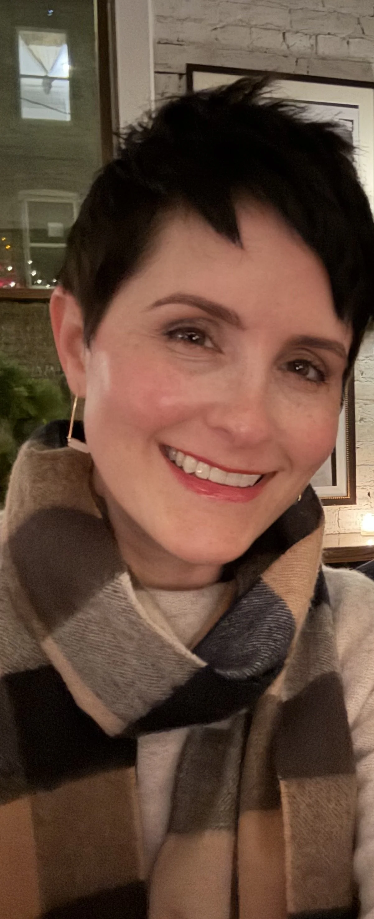 A smiling woman with short black hair, wearing gold hoop earrings and a brown and beige plaid scarf, sitting indoors near a window with white brick walls in the background.