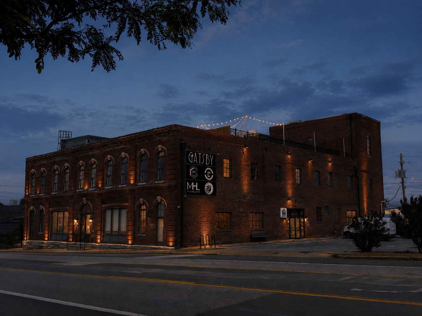 A brick building at dusk with string lights on the rooftop and exterior lighting highlighting its architectural features. The building has large arched windows and a sign reading "GATSBY." There are parked cars and trees in the surrounding area.