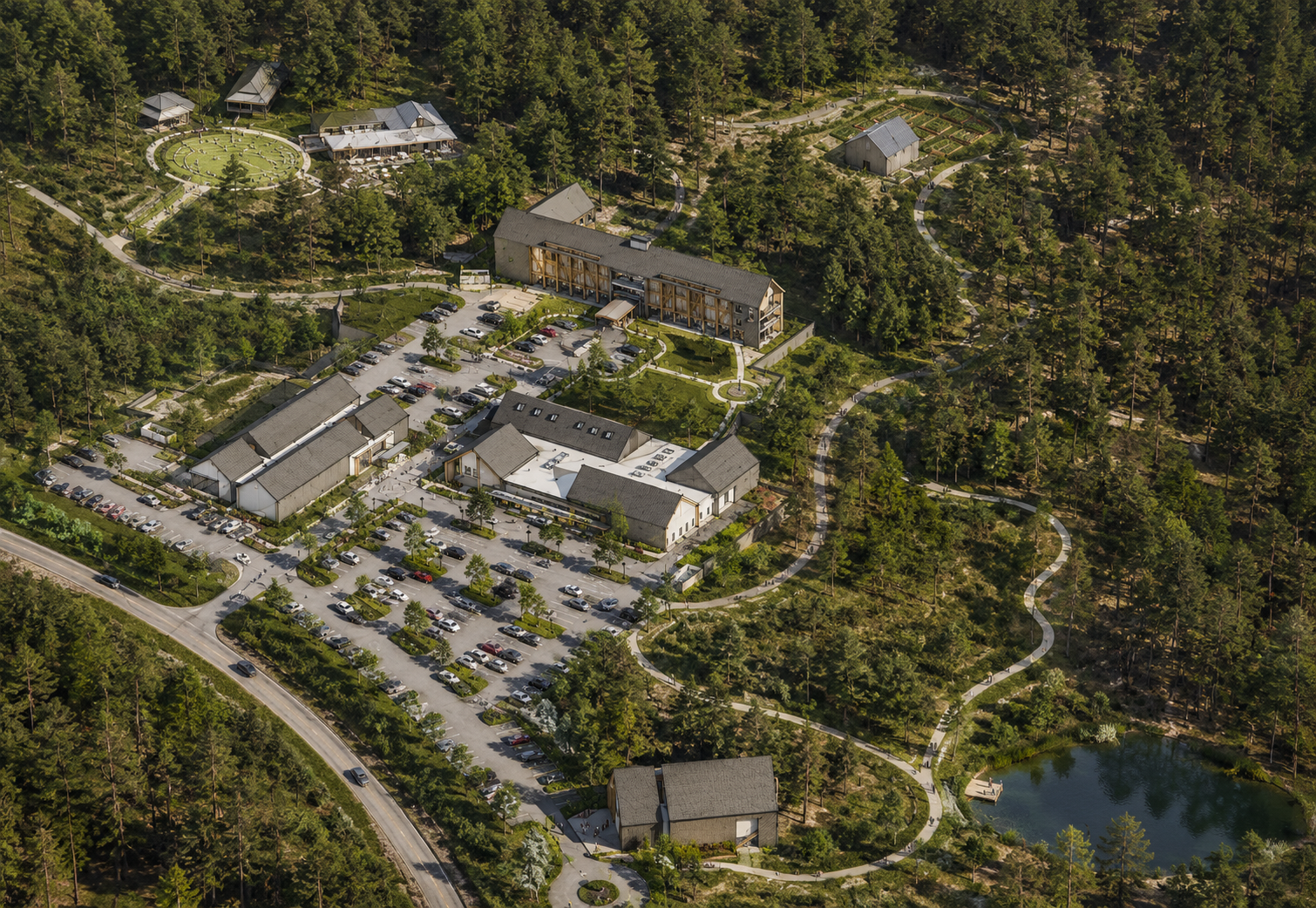 Aerial view of a large, modern building complex surrounded by trees, parking lots, and a winding path leading to a pond, situated in a forested area.