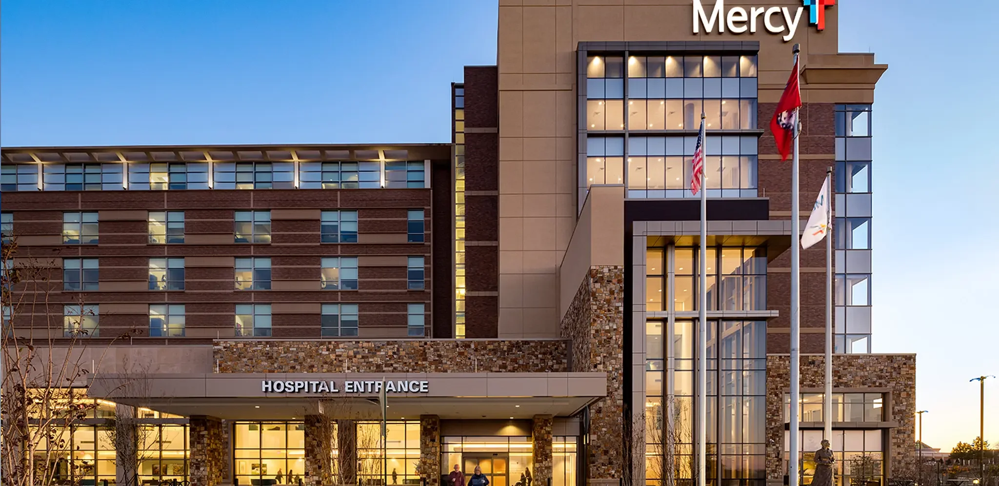 Front view of Mercy hospital with flags in front, lit windows, and a sign that reads 'HOSPITAL ENTRANCE' at dusk.