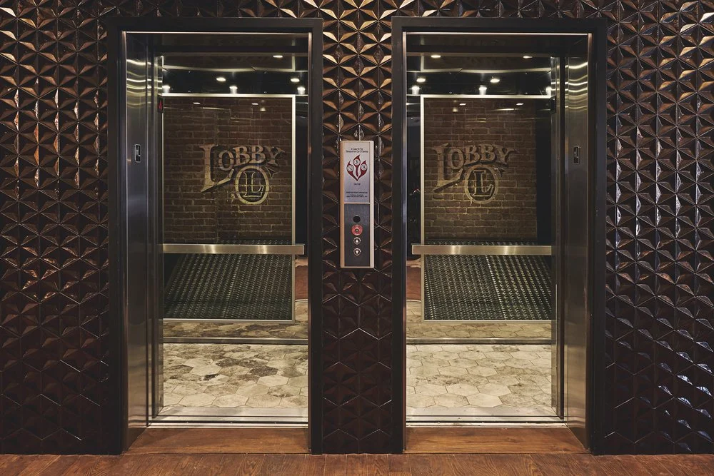 Two glass elevator doors with stainless steel frames, set in a wall with dark geometric patterned tiles. The elevator buttons are in a panel between the doors with a sign above them. The reflection in the glass shows a brick wall with the word "Lobby" written on it.
