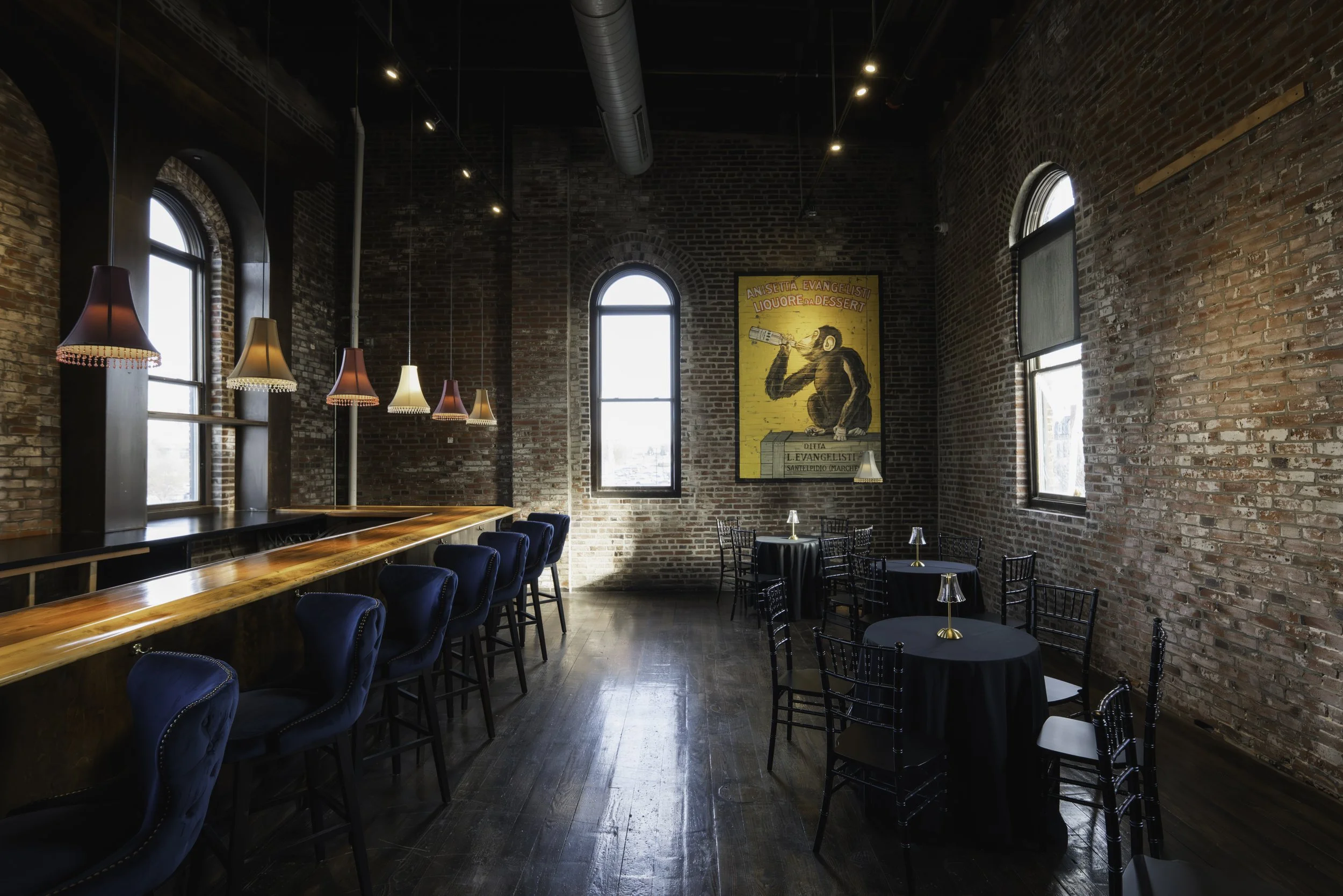Interior of a rustic brick-walled cafe or restaurant with large arched windows, hanging pendant lights, and small round tables with chairs.