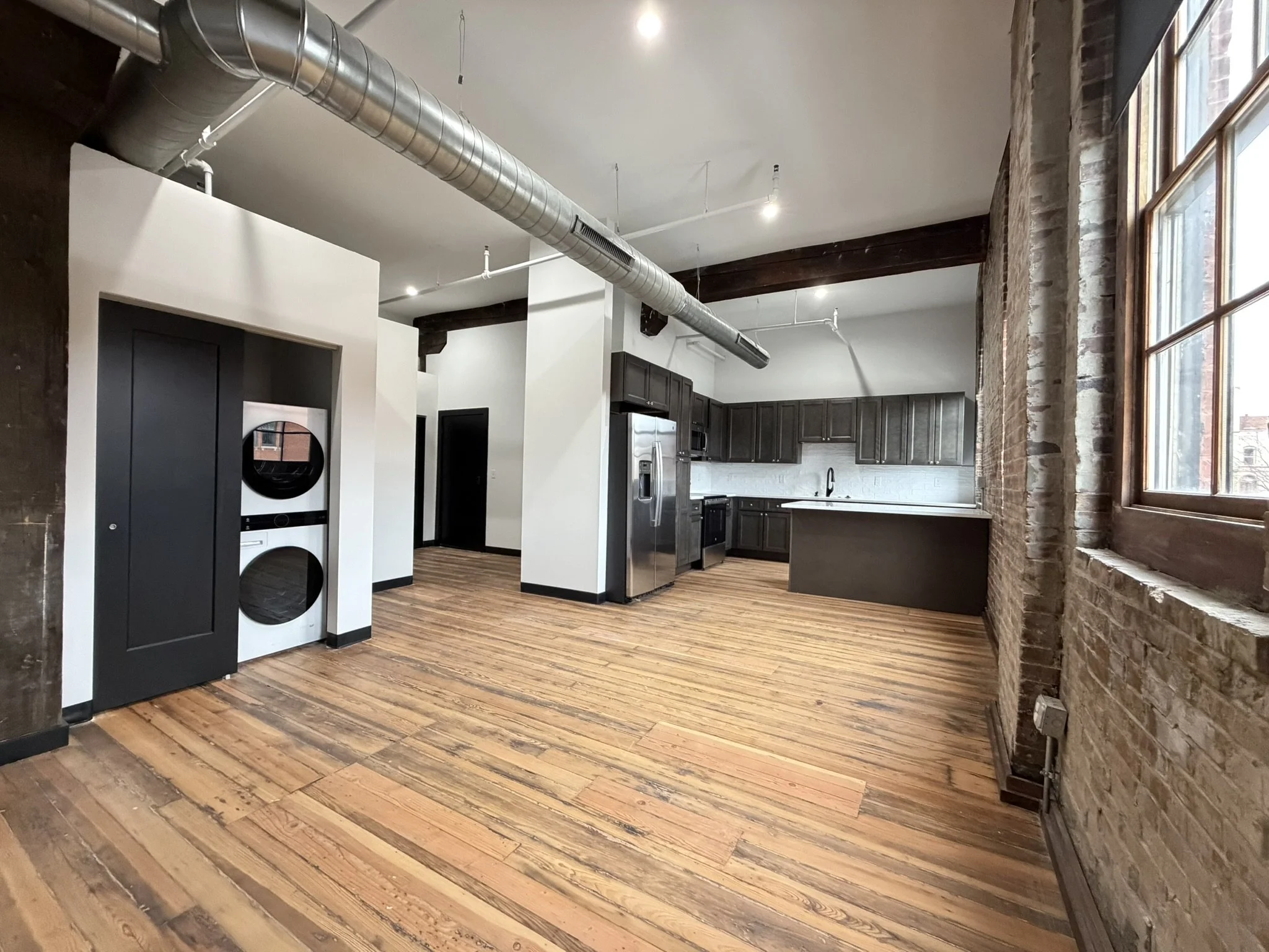 Interior of a modern apartment with exposed brick walls and large windows, showing kitchen cabinets, stainless steel appliances, hardwood floors, and visible ductwork on the ceiling.