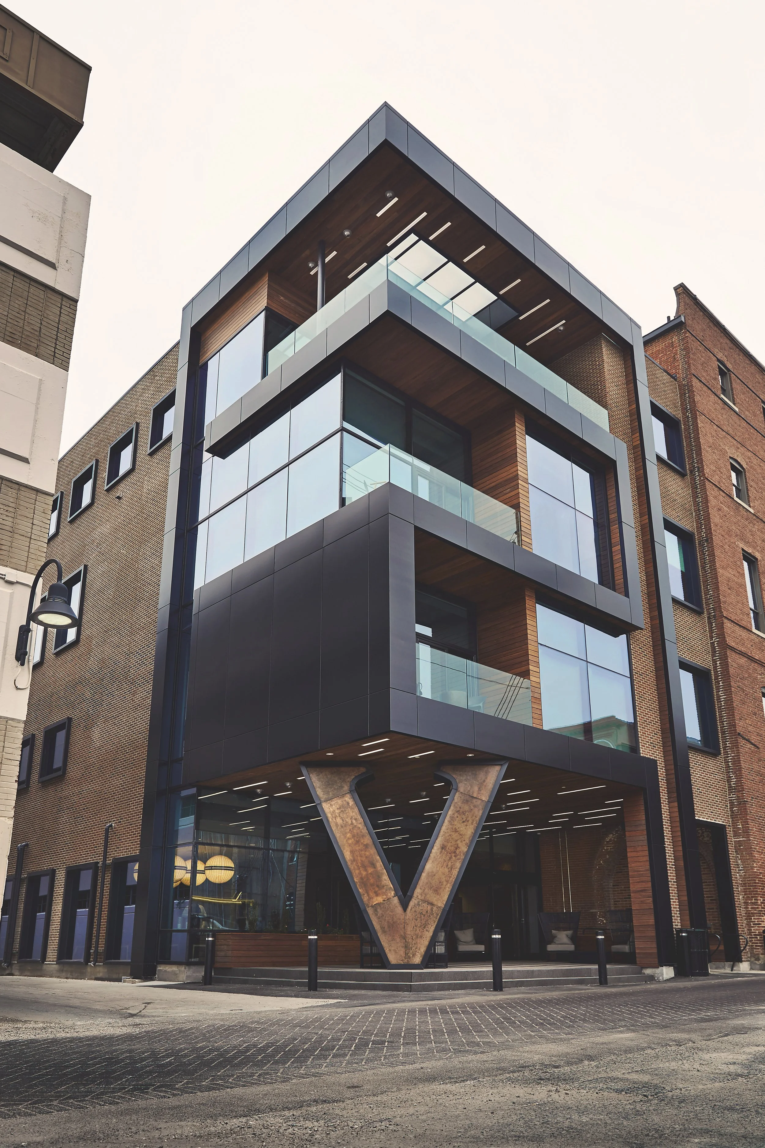 Modern multi-story building with glass balconies and a prominent V-shaped support structure at the entrance, with neighboring brick buildings and street lamp in an urban setting.