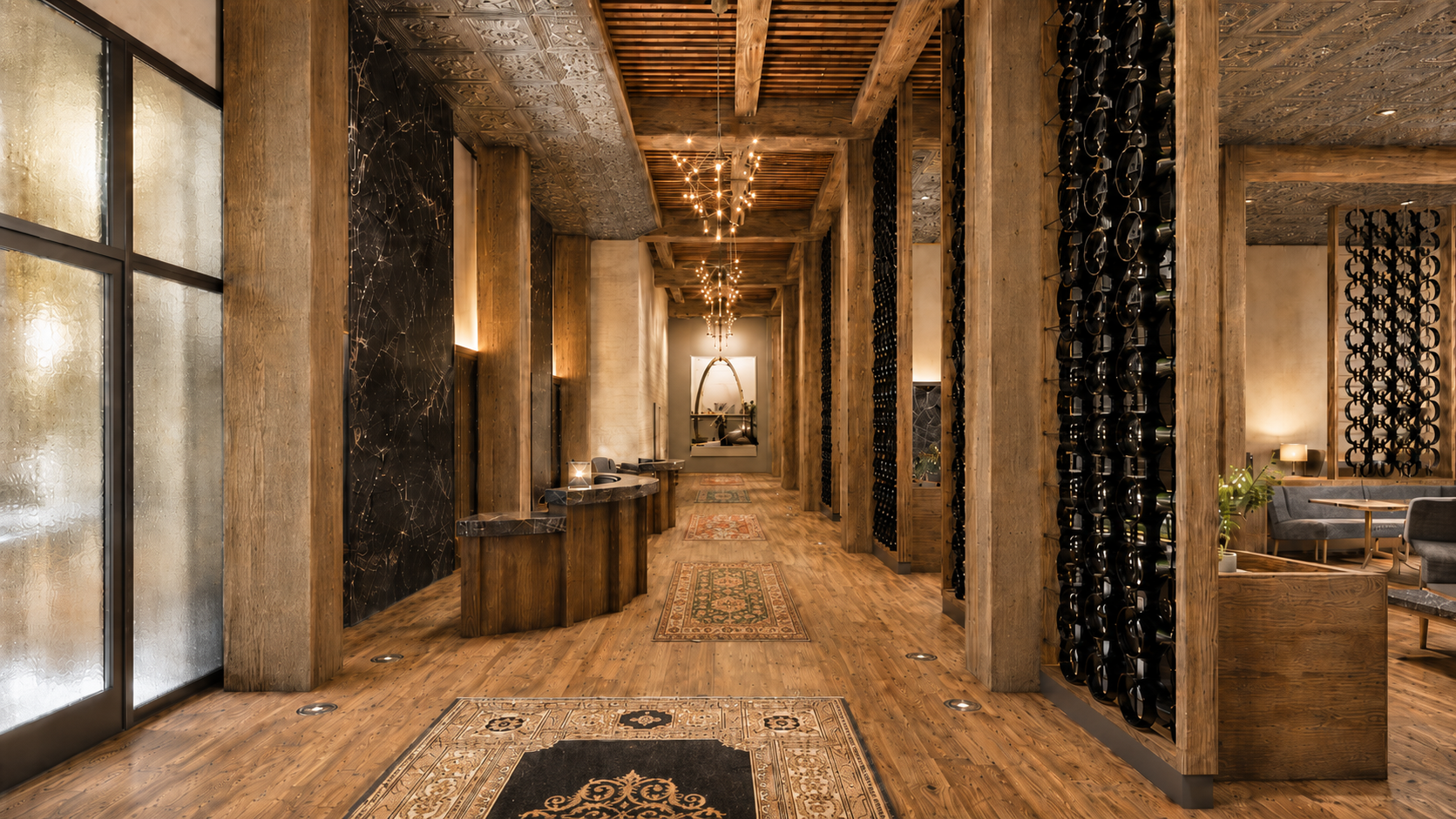Interior of a cozy, upscale restaurant or hotel lobby with wooden floors, walls, and ceiling beams. Dim lighting, decorative rugs, and a unique lighting fixture hanging from the ceiling. Black wine bottles are stored on racks on the wooden beams, and there's a sitting area with tables and chairs to the right.