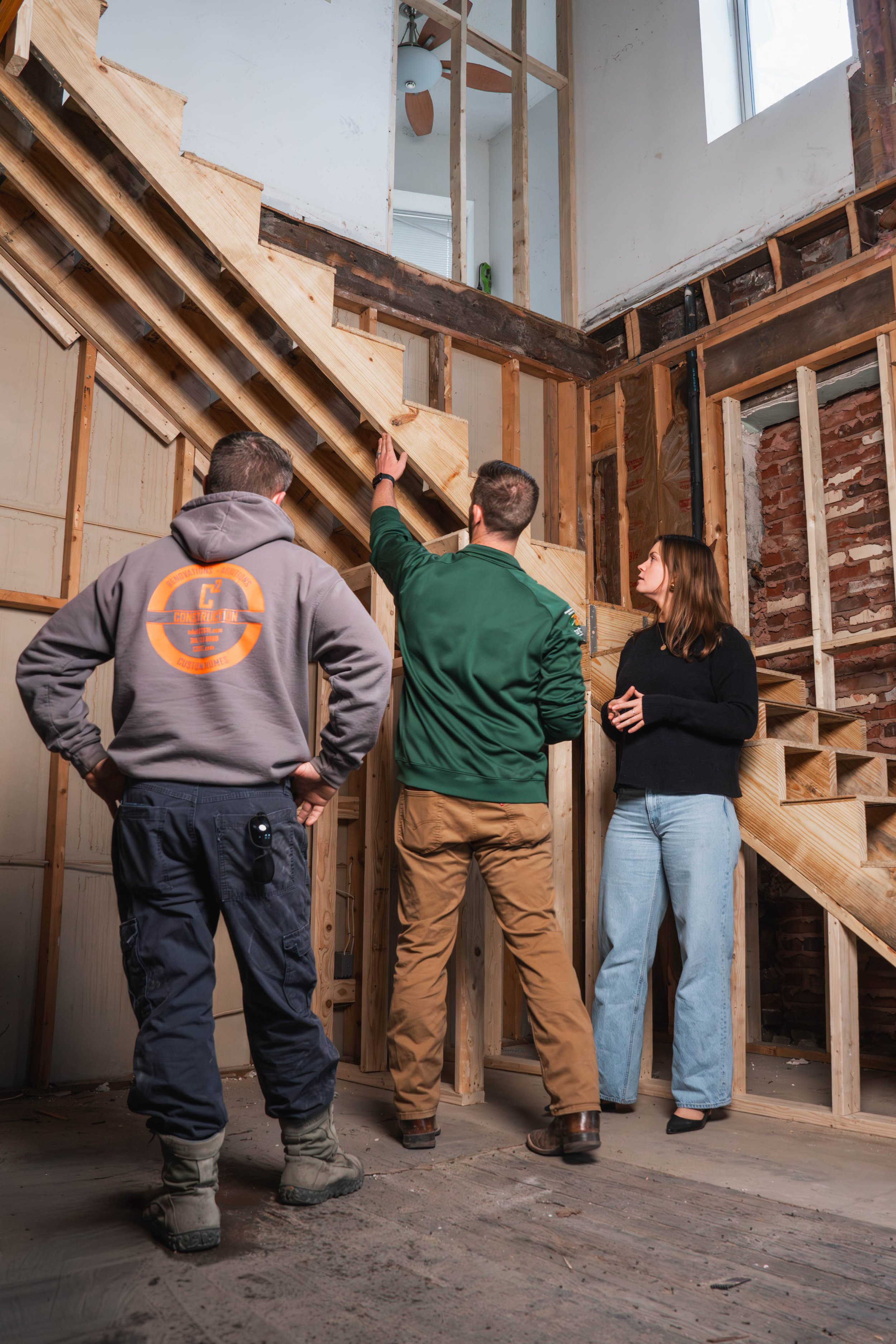 Three people are inside a house under construction, with exposed wooden framing and insulation. They are discussing the ongoing construction work, with one person pointing towards the stairs.