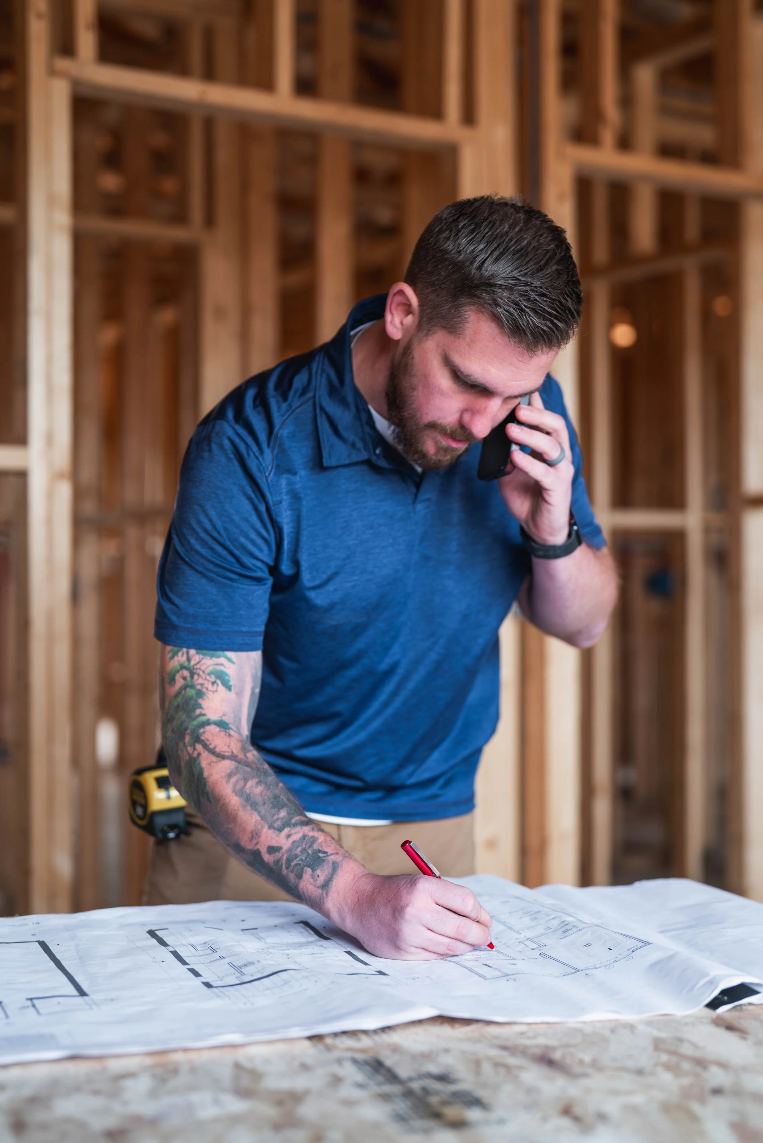 Man with tattoos wearing a blue shirt and beige pants, talking on a phone while examining architectural plans on a construction site with wooden framing in the background.