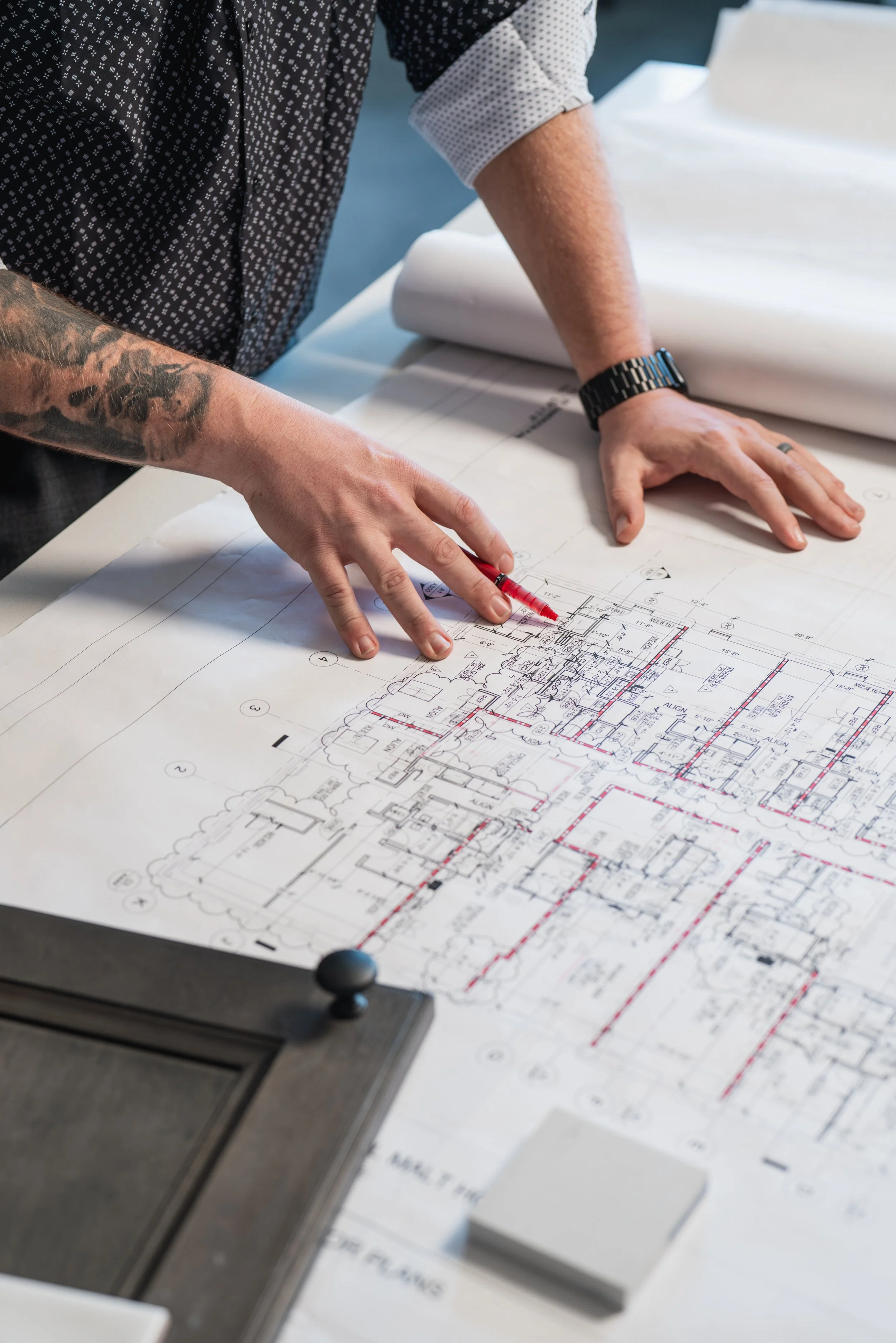 A person is working on architectural blueprints spread out on a table. They are using a red pen and are wearing a watch. Part of a drafting machine is visible on the left side of the table.