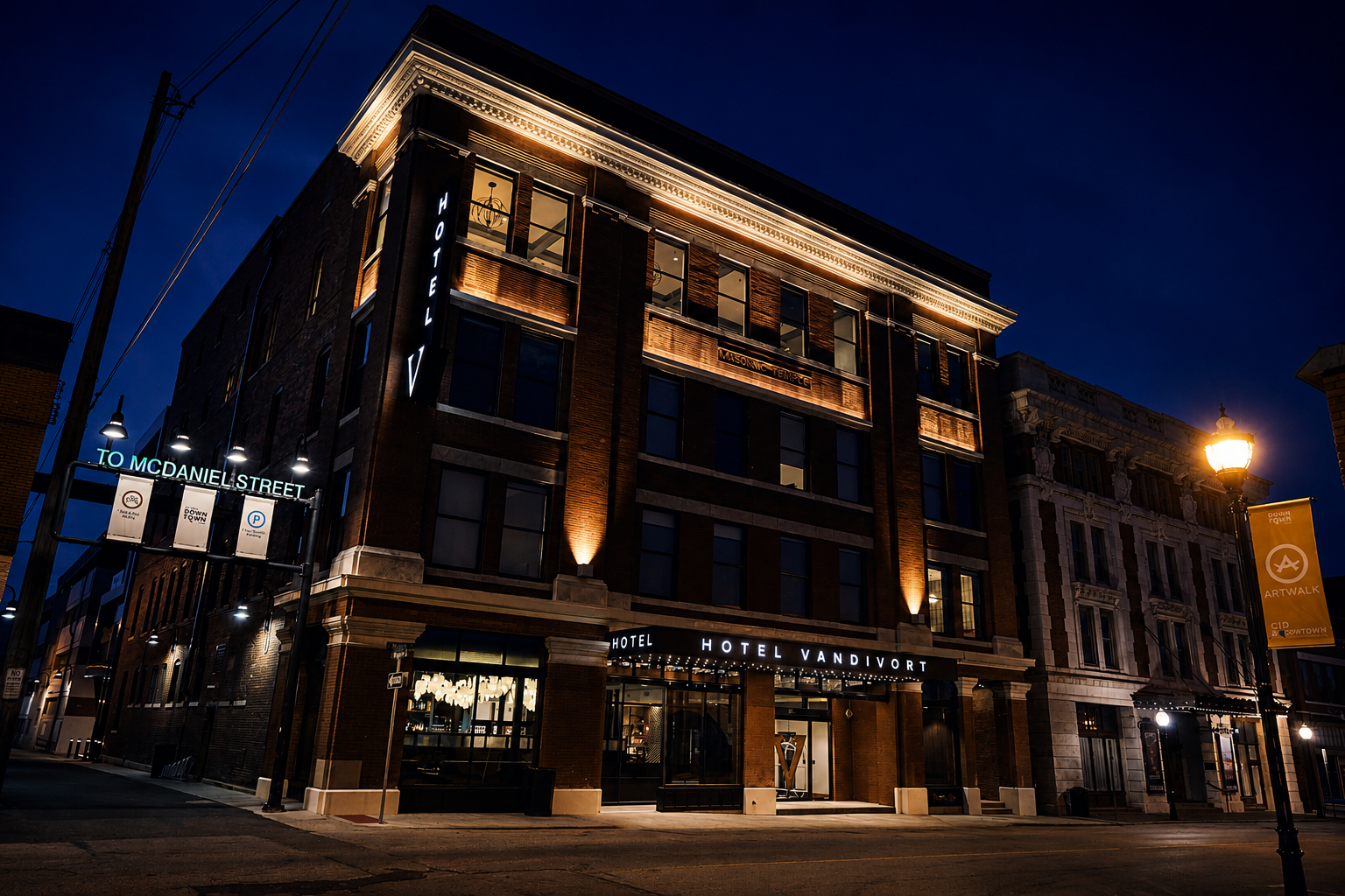 Nighttime view of a brick hotel named Hotel Vandivort with illuminated signage. The building features large windows, decorative lighting, and is located on a city street with a streetlamp and banners.
