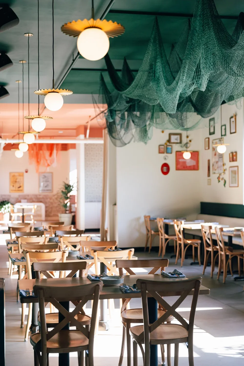 Empty dining area with wooden tables and chairs, hanging light fixtures, decorative ceiling nets, and framed pictures on the wall.