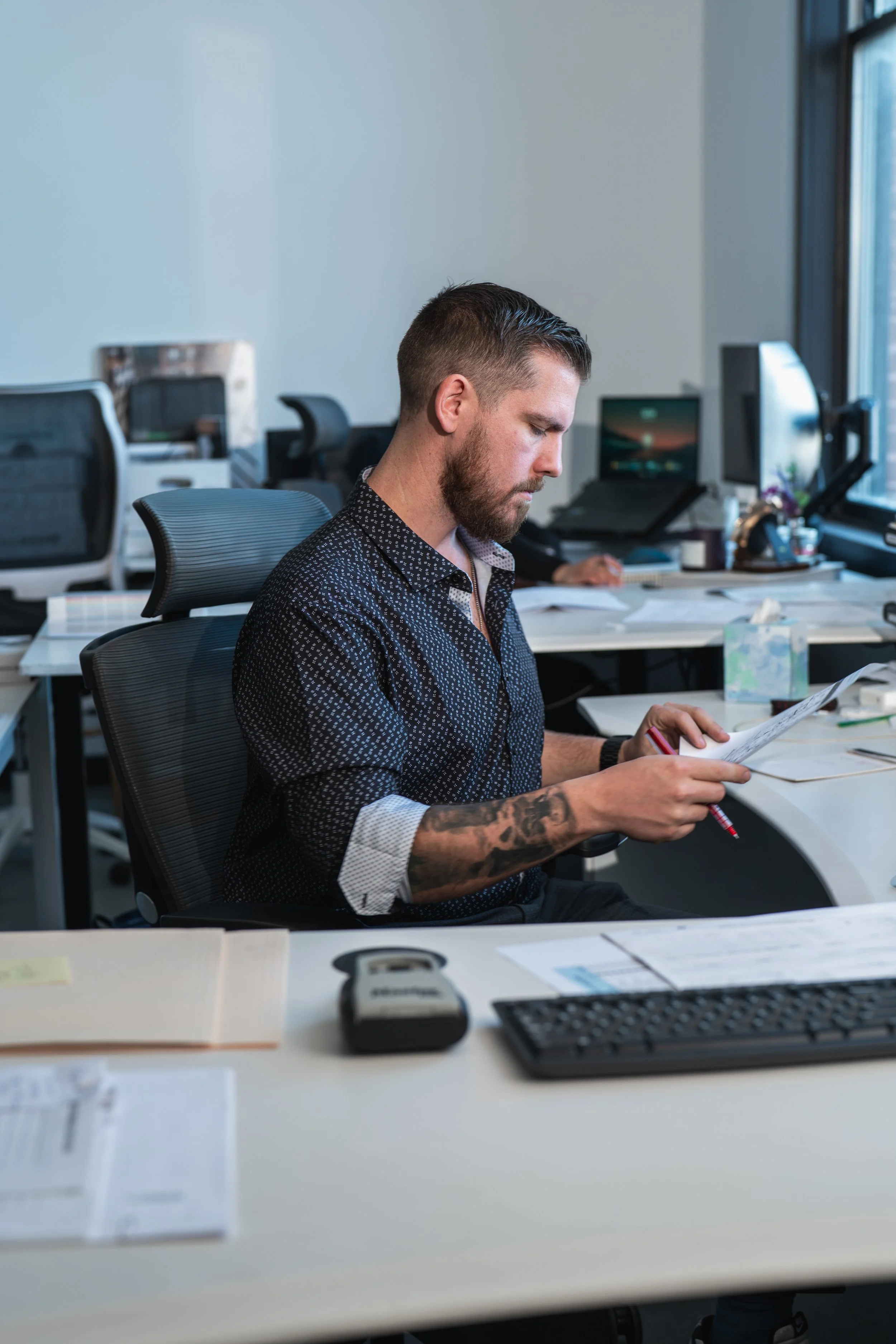 A man with a beard, tattooed arm, and short hair, wearing a black patterned shirt, is sitting at an office desk looking at a sheet of paper with a pen in his hand. The office has multiple desks, chairs, a computer monitor, and a window.